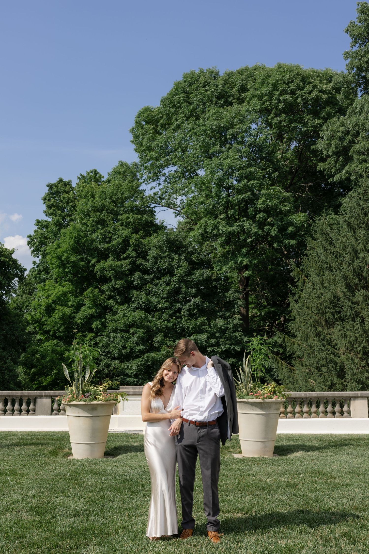 Couple embracing on a grassy lawn with decorative planters and trees in background.