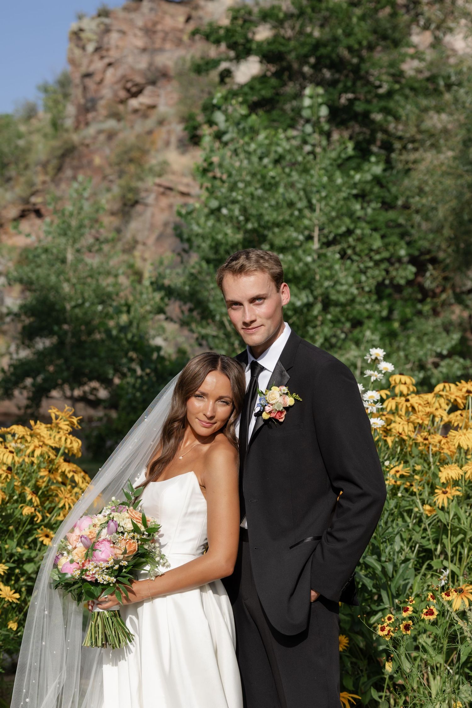 Bride and groom pose together in garden with yellow flowers and mountain backdrop.