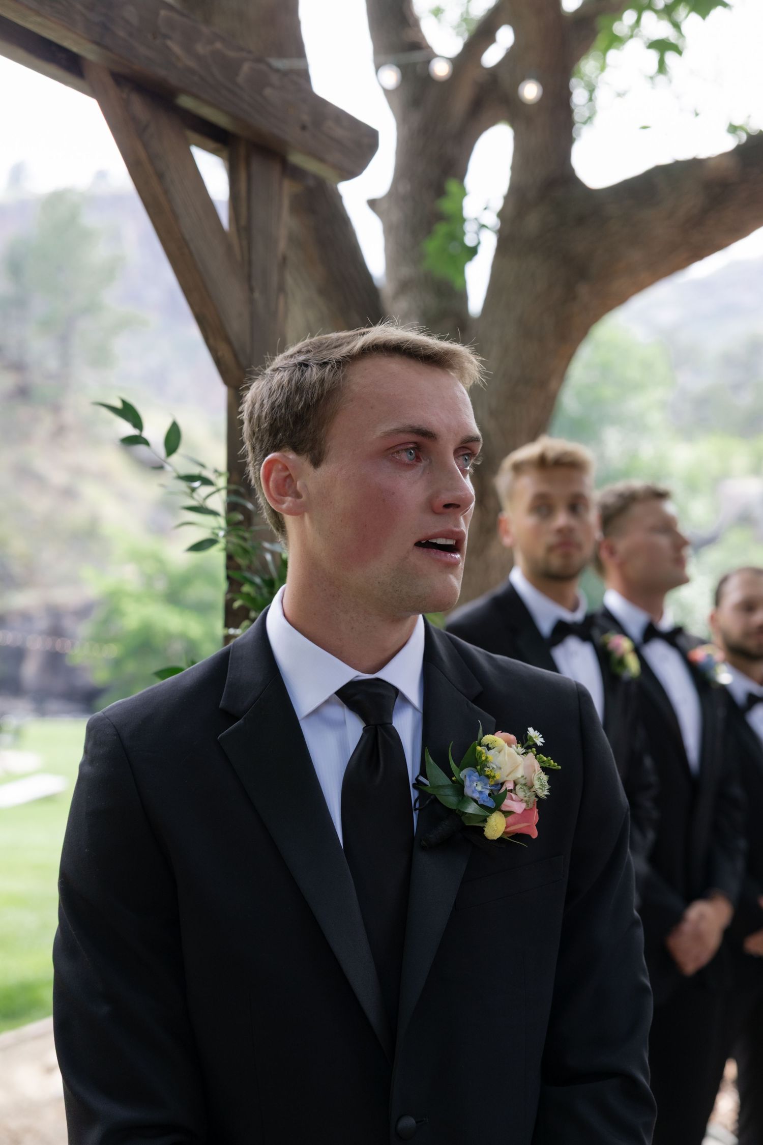 Groomsmen in black suits and ties at an outdoor wedding ceremony.