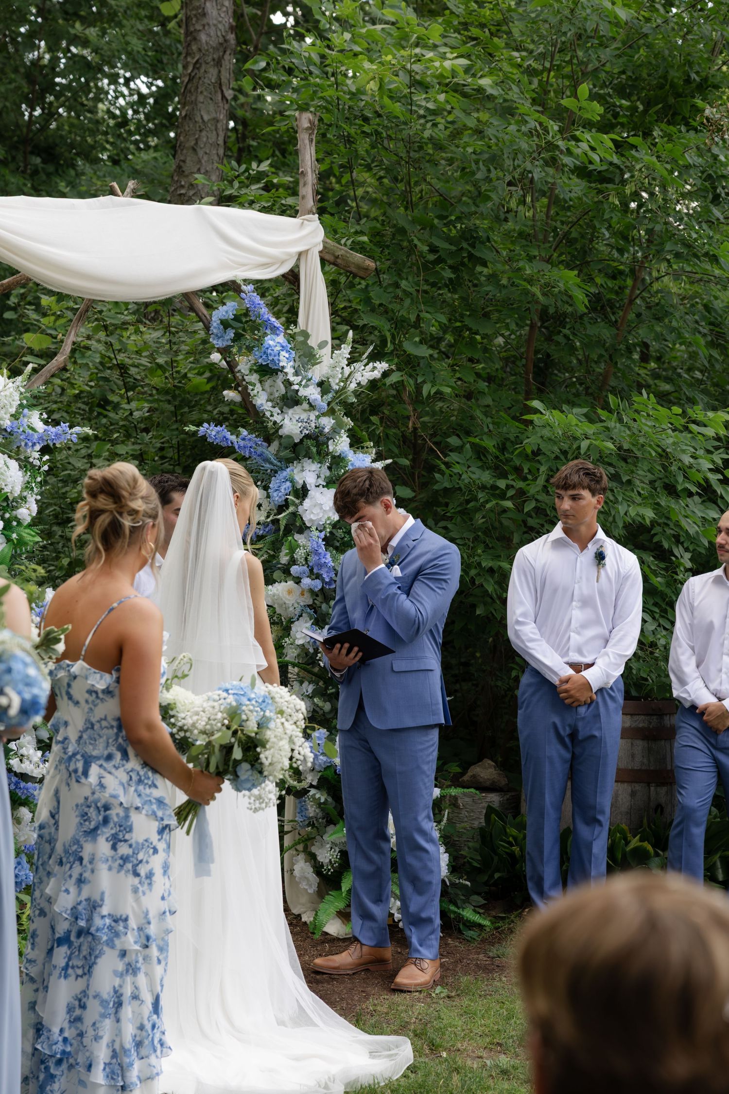 Wedding party in blue suits stands beneath a white fabric arch decorated with blue and white flowers.