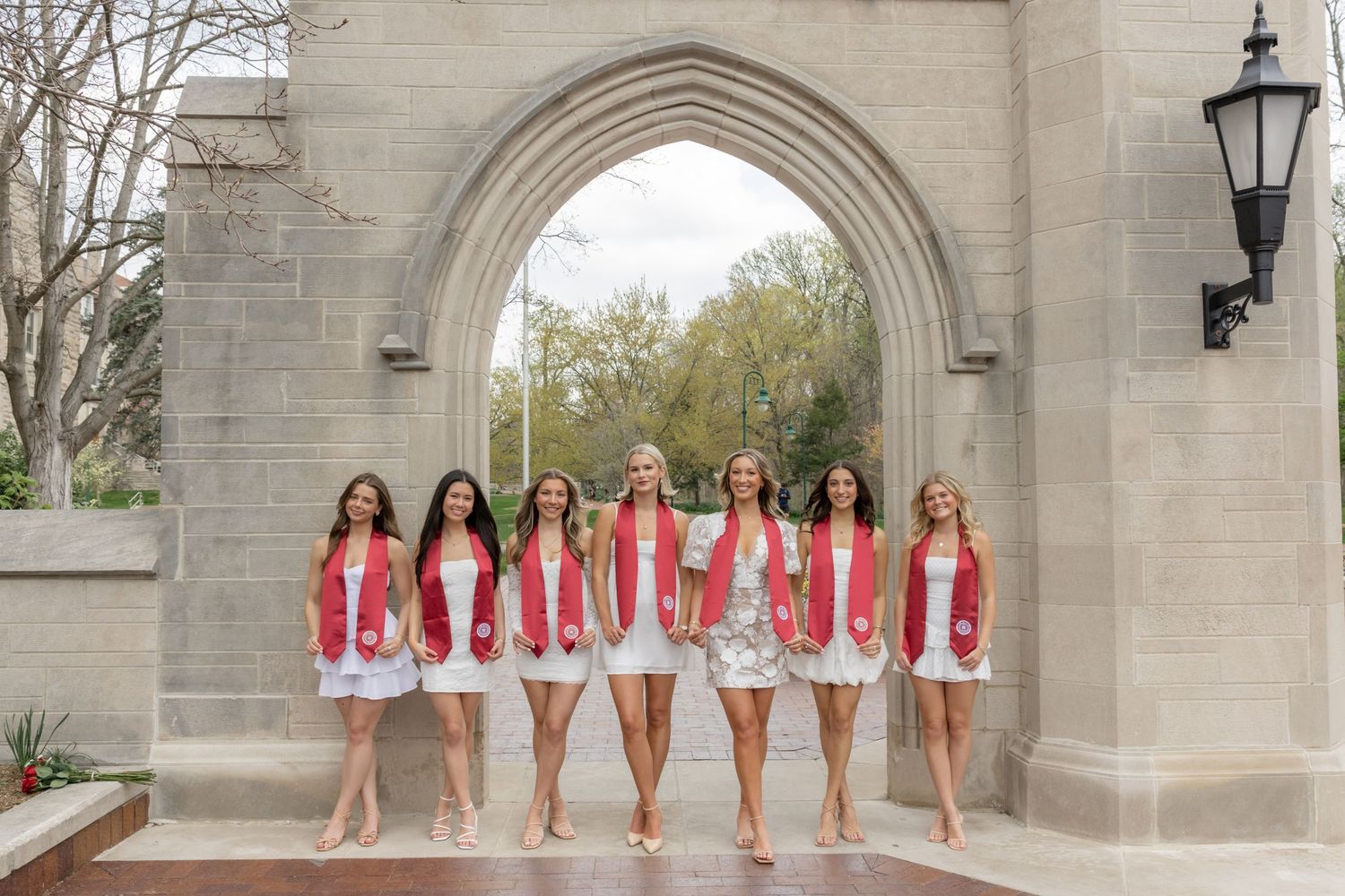 Group poses in matching white dresses and red sashes under stone archway during graduation photoshoot.