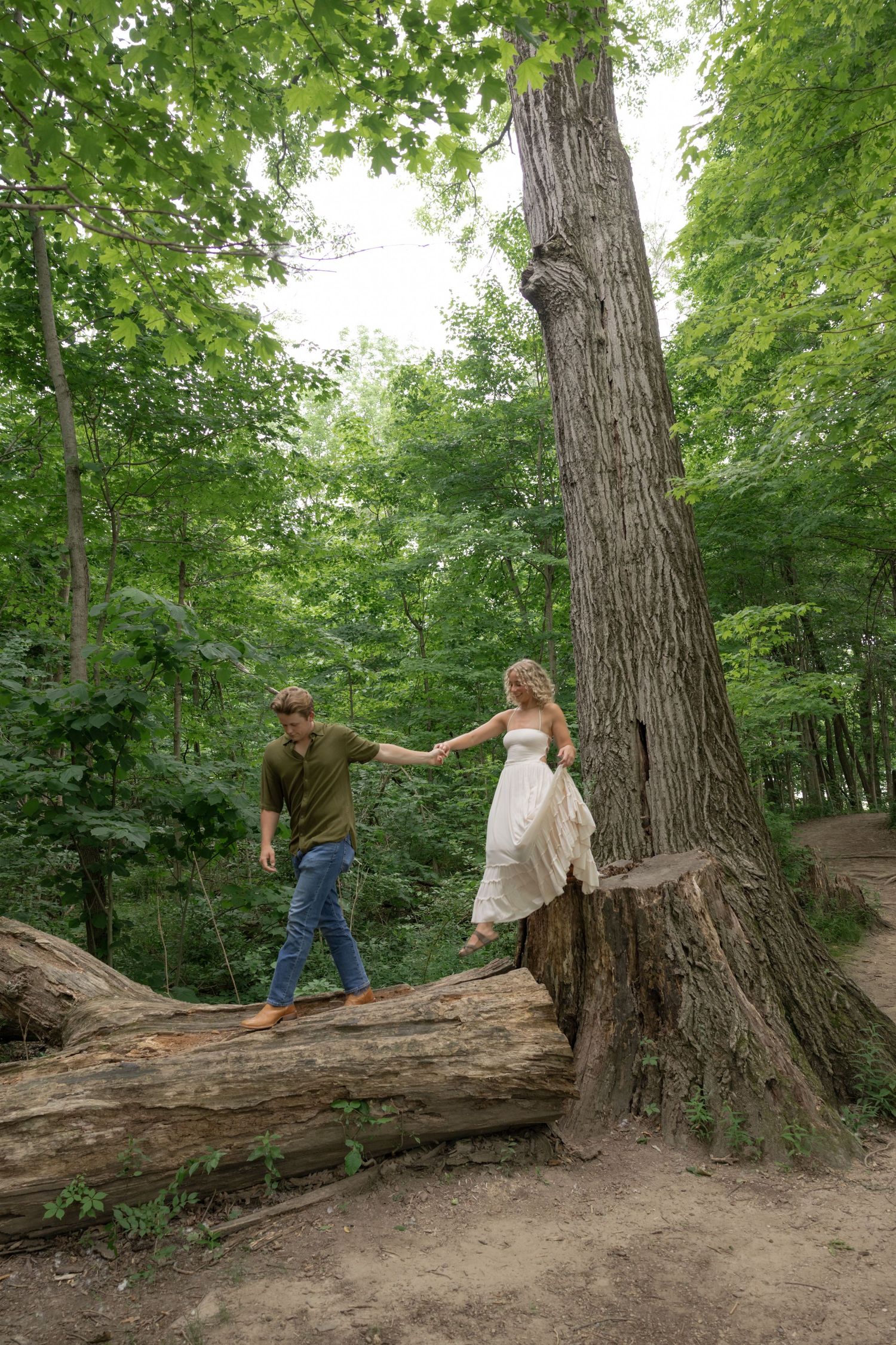 A couple walks and poses playfully on a large fallen tree trunk in a lush green forest.