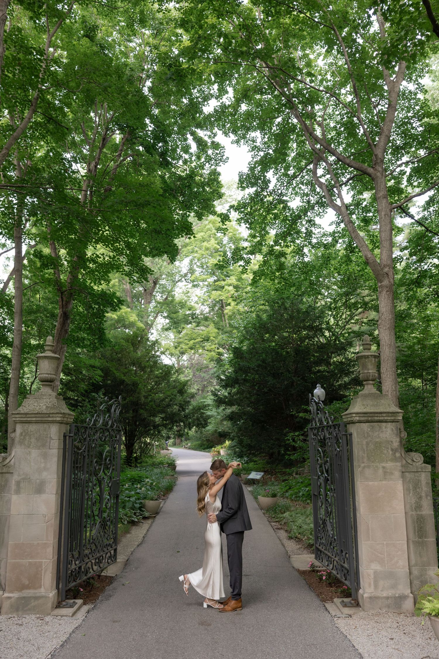 A tree-lined pathway with stone pillars and iron gates frames a couple in the distance.