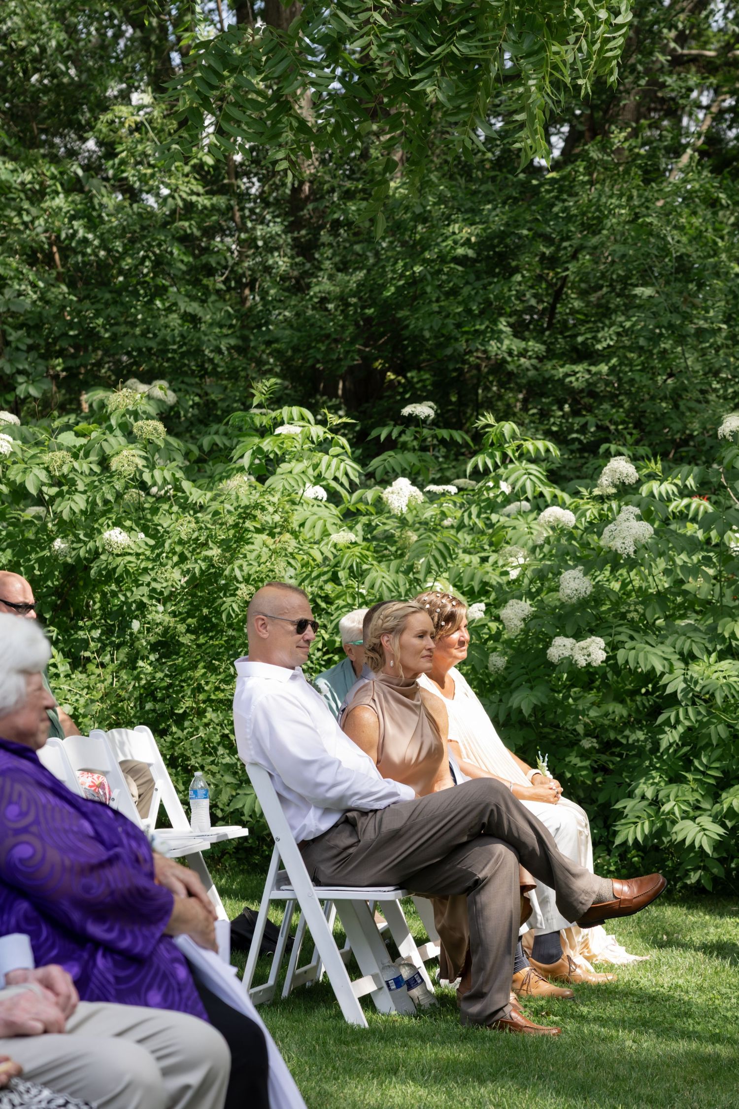 Wedding guests seated on white chairs in a lush garden setting during an outdoor ceremony.