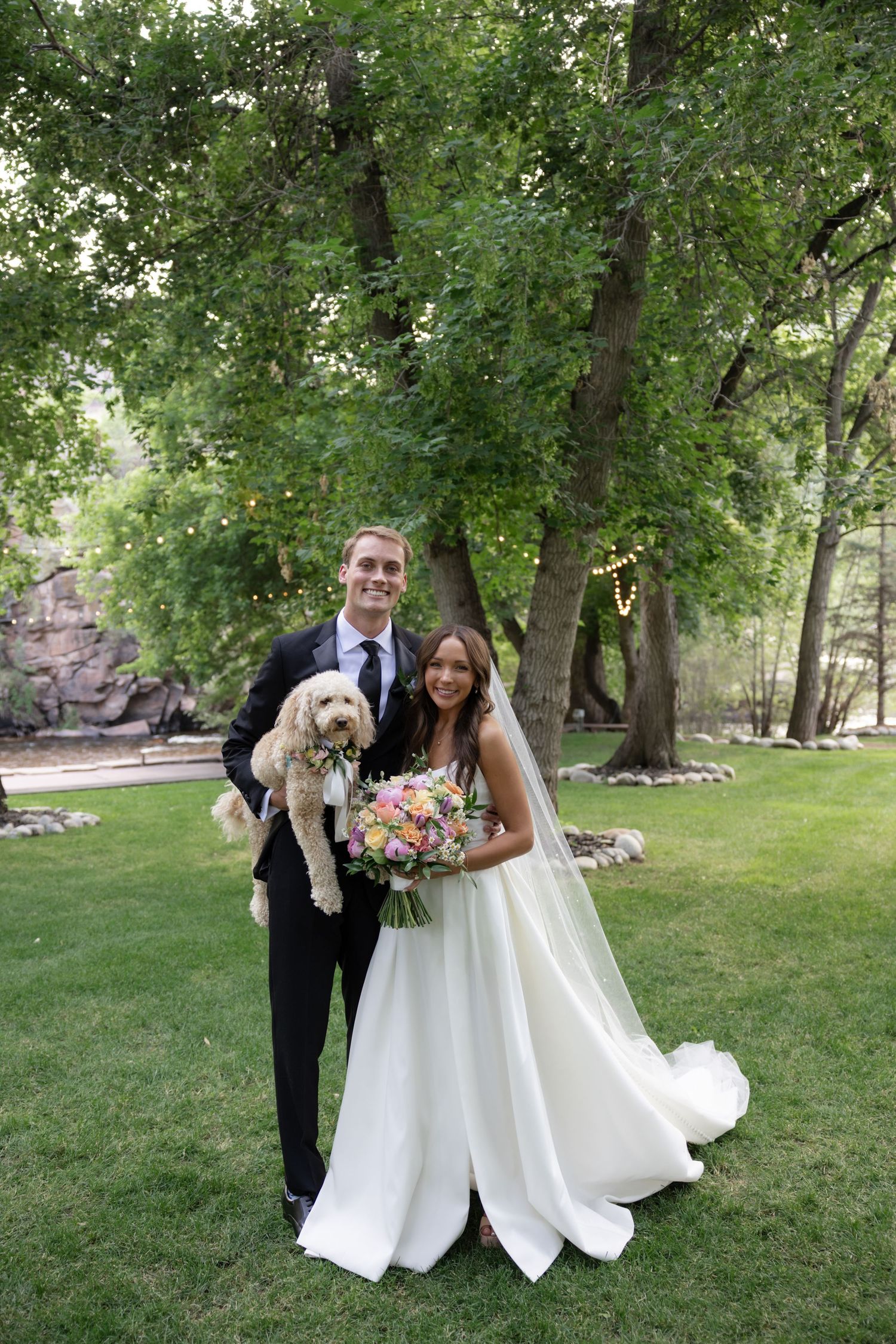 Wedding couple poses with white fluffy dog in outdoor garden setting.