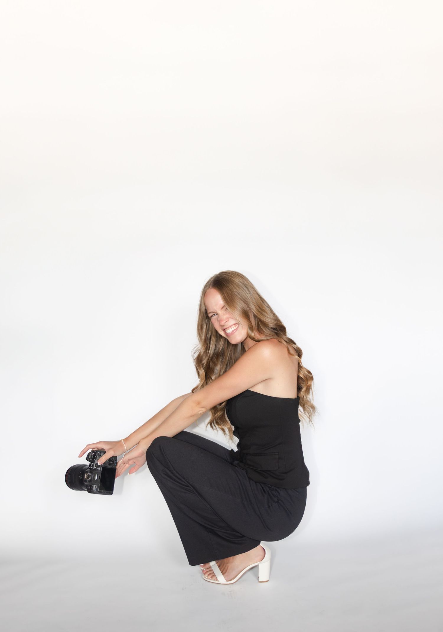 Photographer in black outfit crouches down while holding camera in studio setting.
