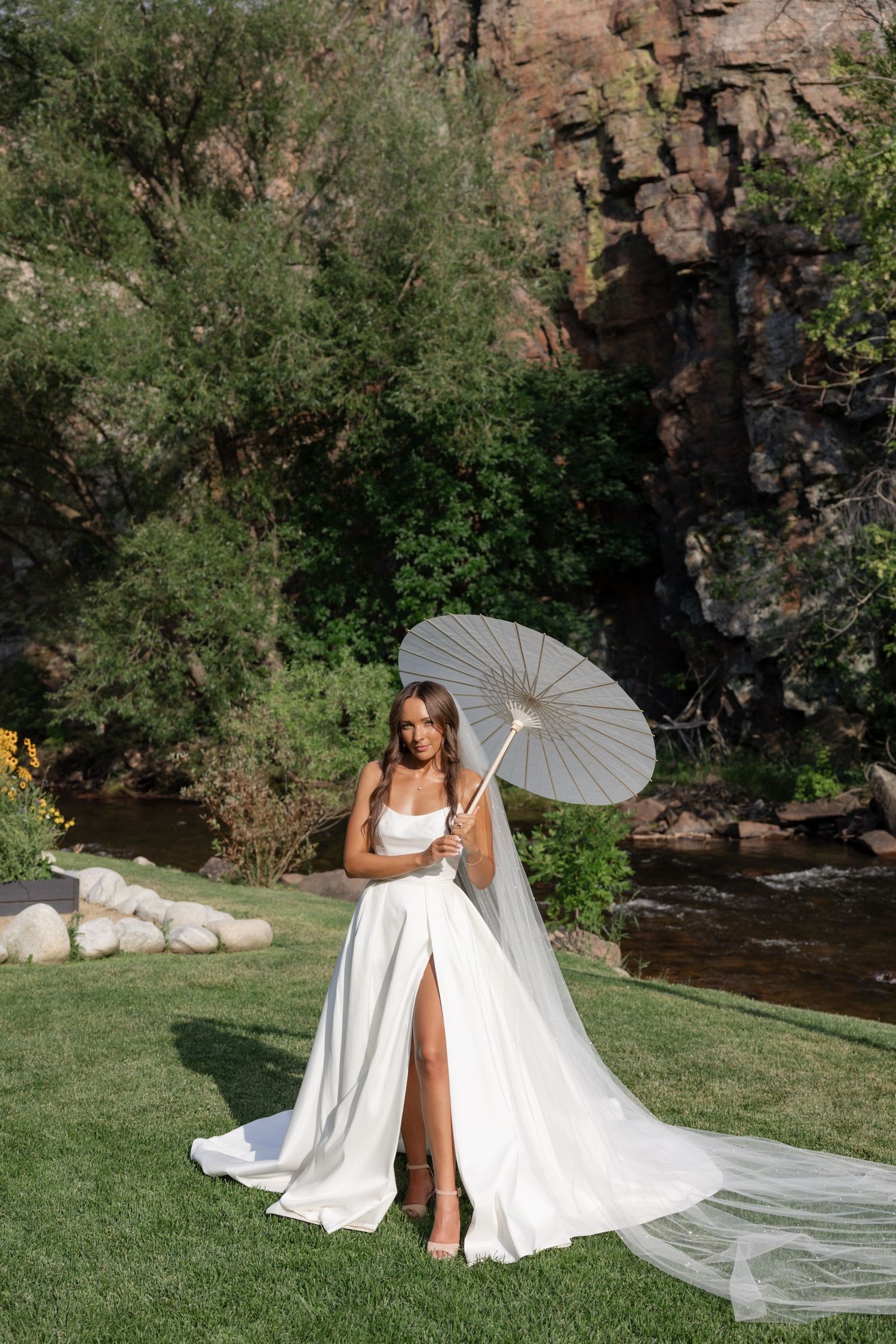 Bride in flowing white dress poses gracefully with parasol against rocky mountain backdrop.