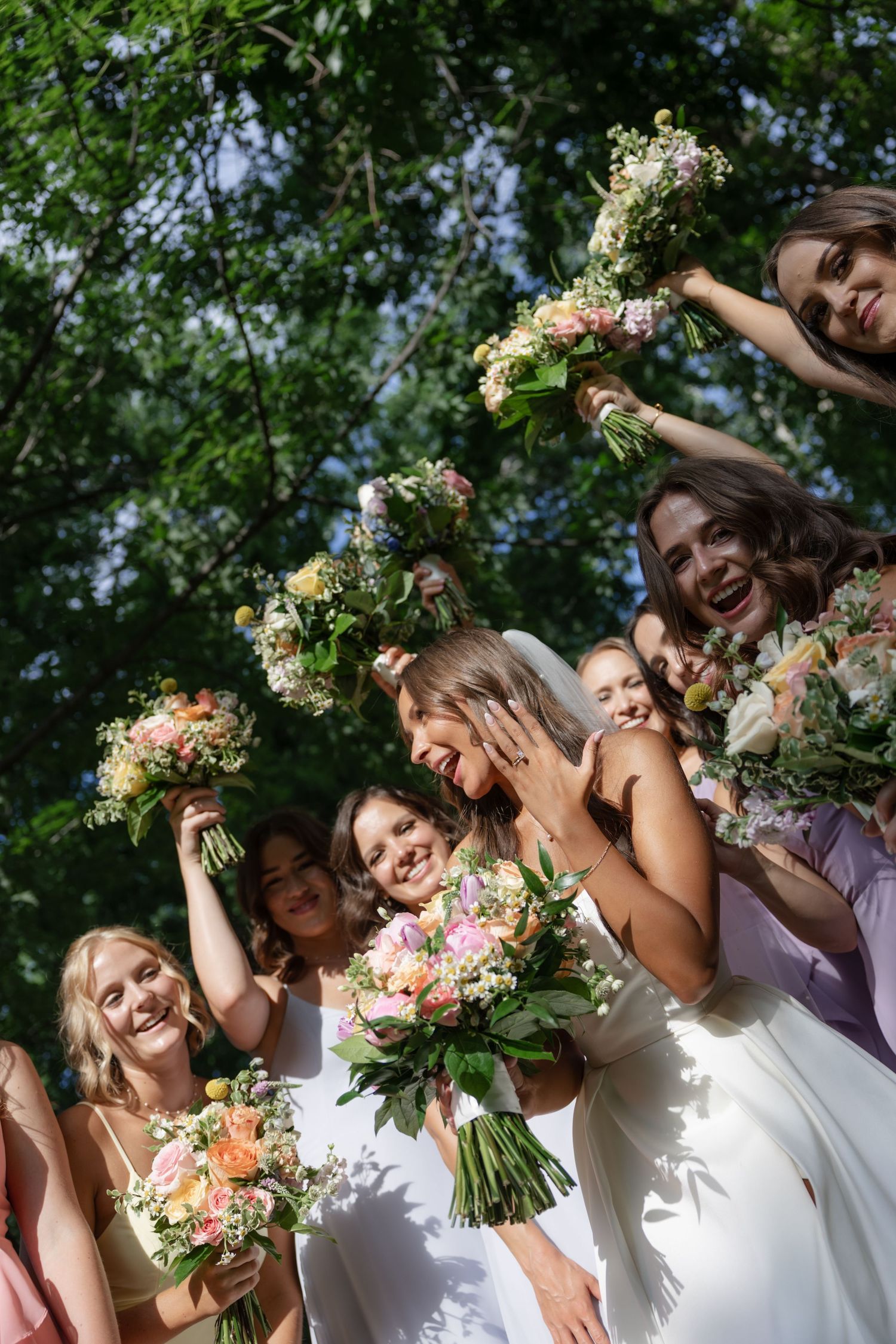 Bridesmaids in white dresses pose with colorful bouquets under trees at an outdoor wedding.