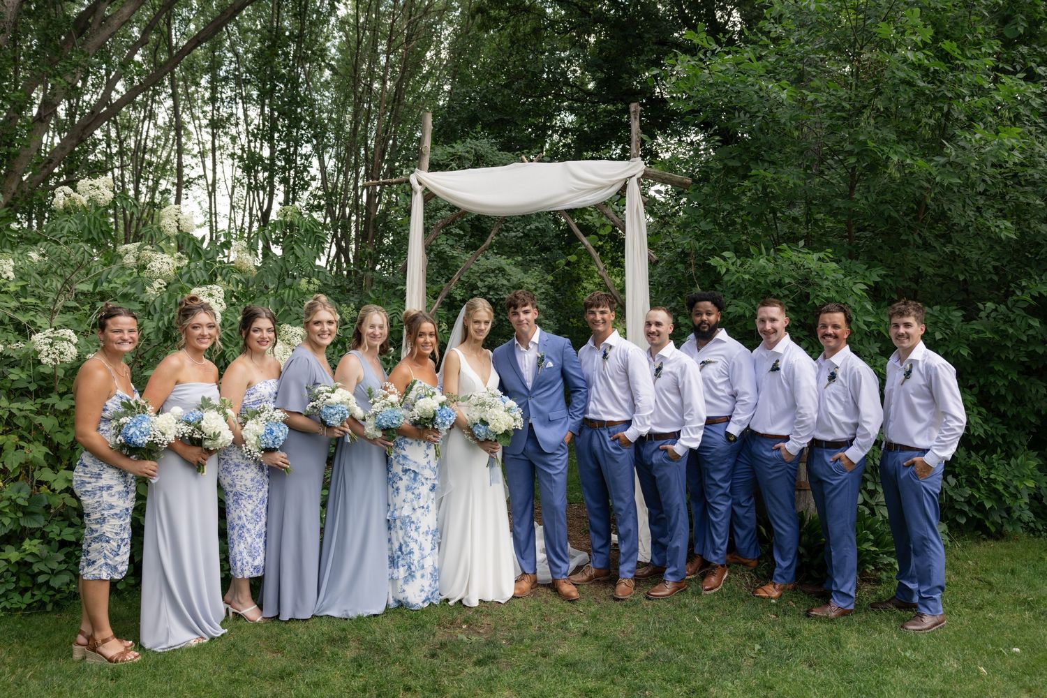 Wedding party in coordinated blue and gray outfits standing together for formal group photos.