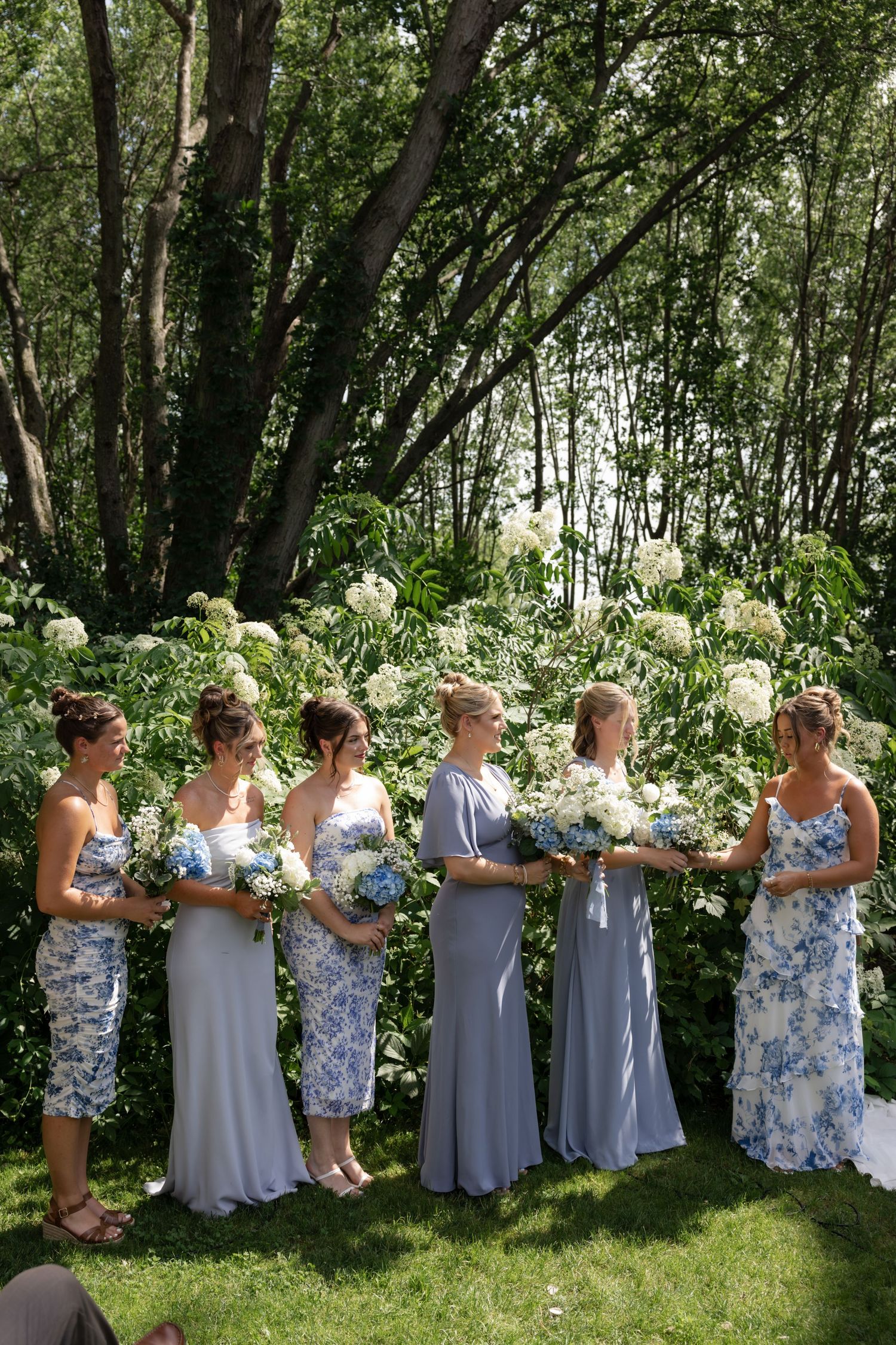 Bridesmaids in blue floral and gray dresses stand together holding bouquets in a wooded garden setting.