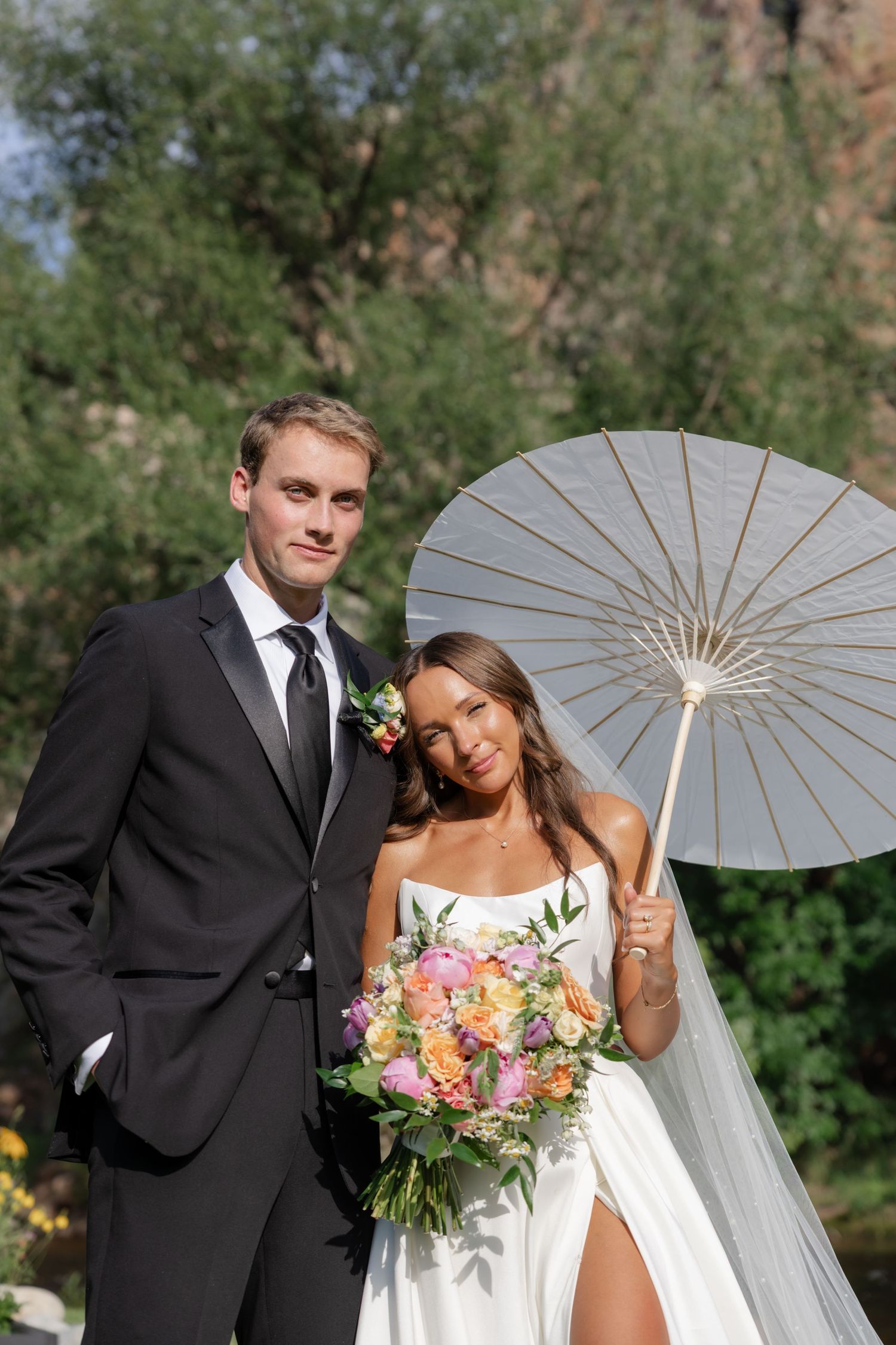 Wedding couple poses with decorative white parasol in garden setting.