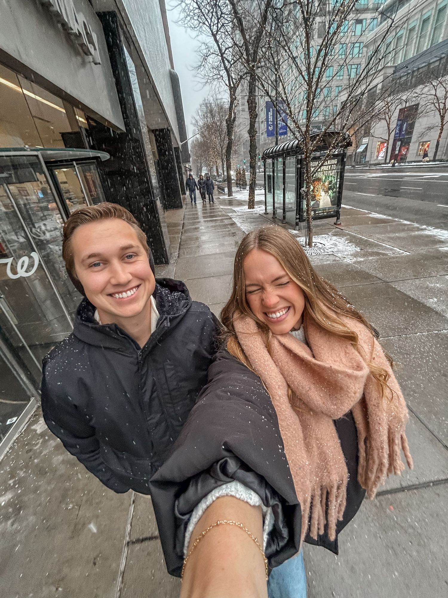 People taking selfies on a snowy city street with tall buildings in the background.