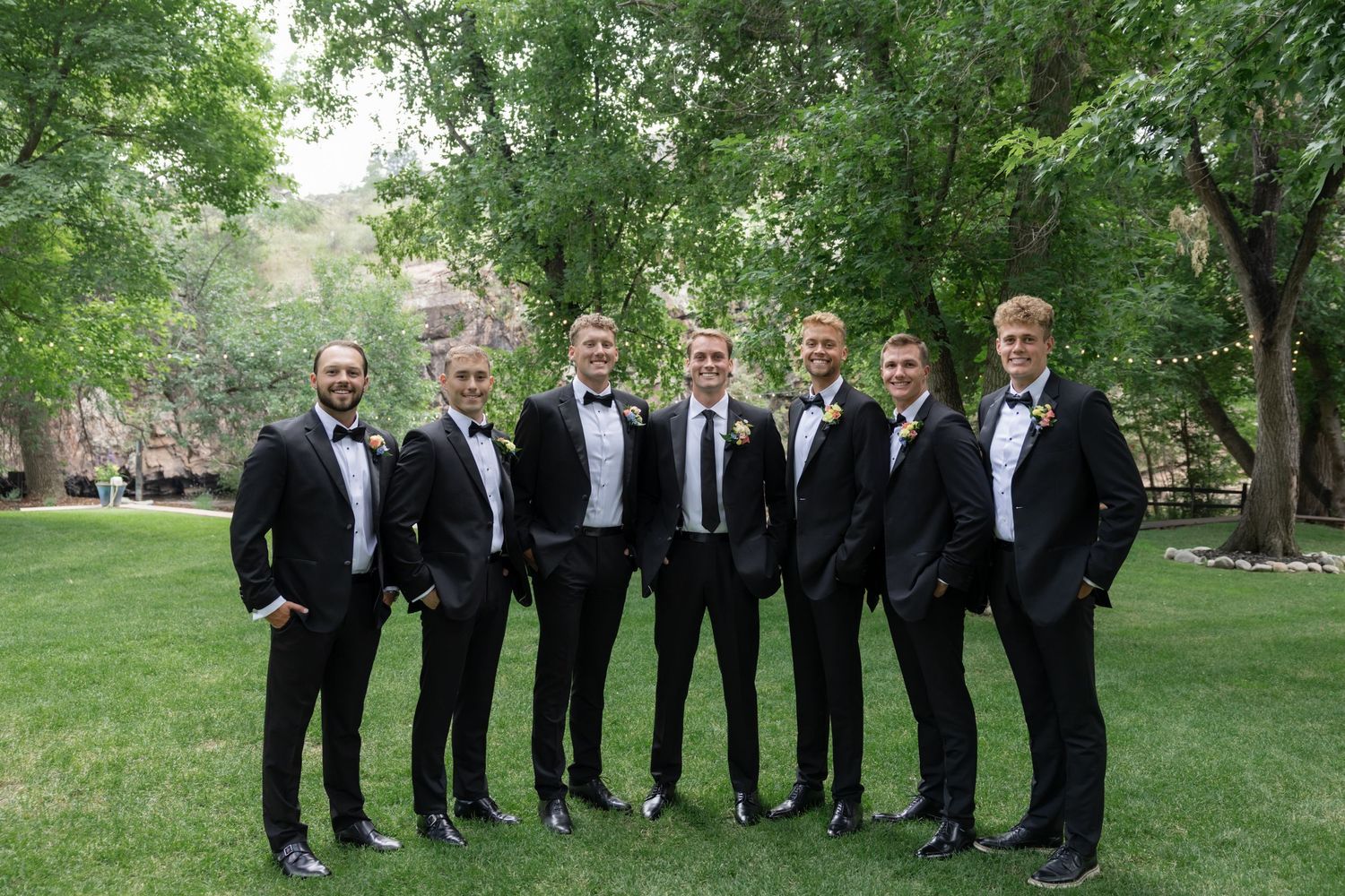 Groomsmen in black tuxedos lined up for a formal wedding photo in a garden setting.