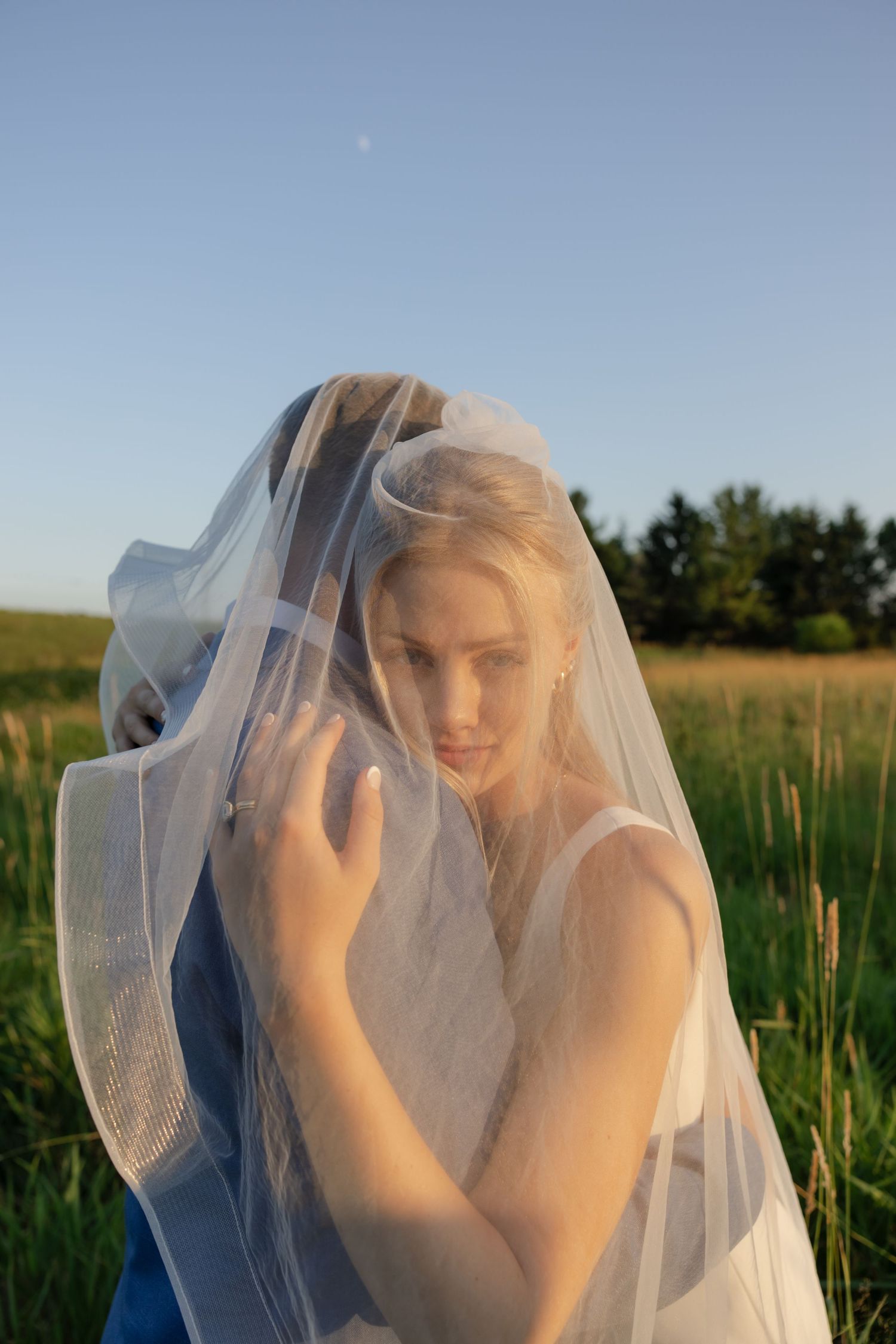 Artistic portrait series of a bride shrouded in delicate white veil against a blue sky and field.