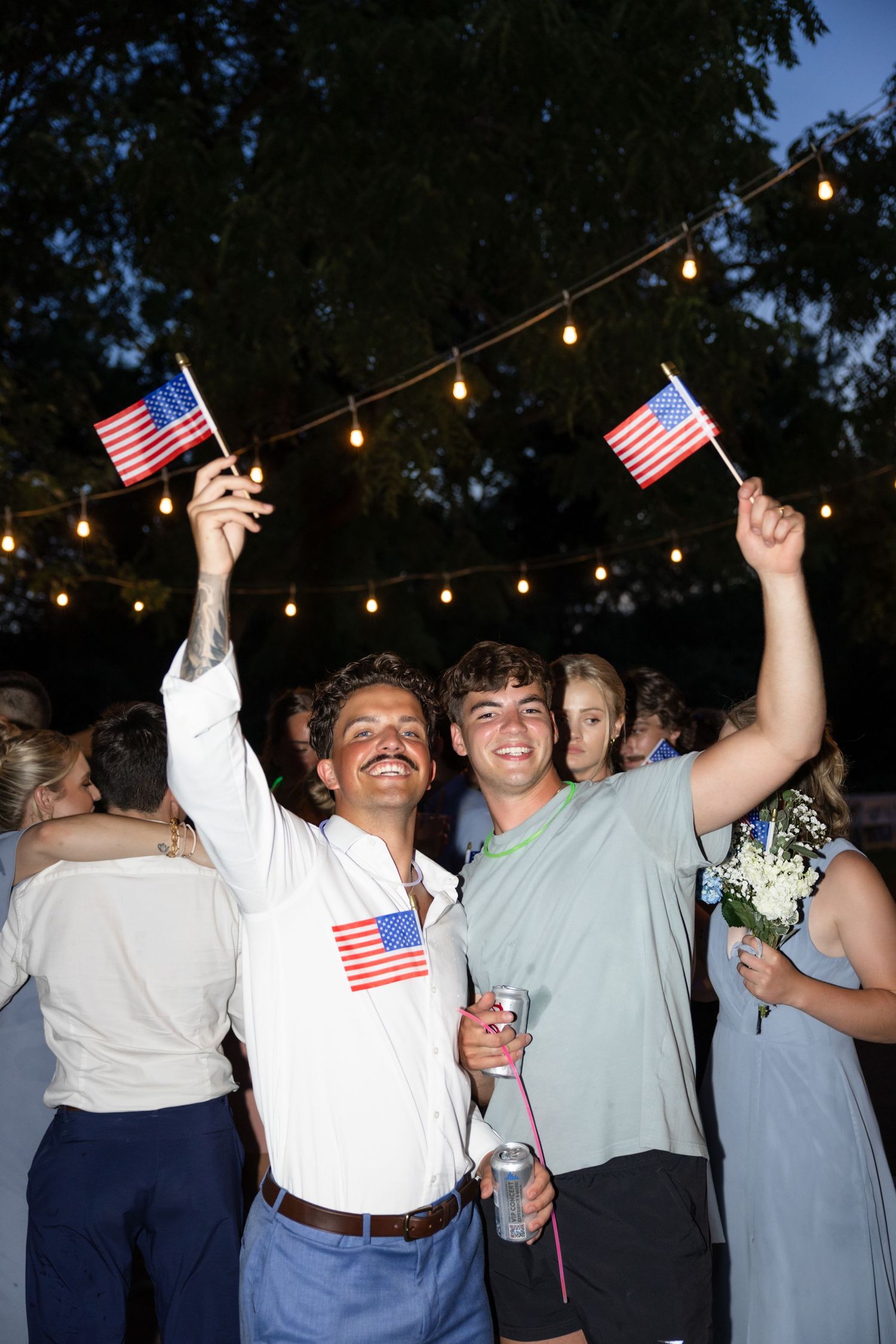 Celebratory crowd holds American flags under outdoor string lights.