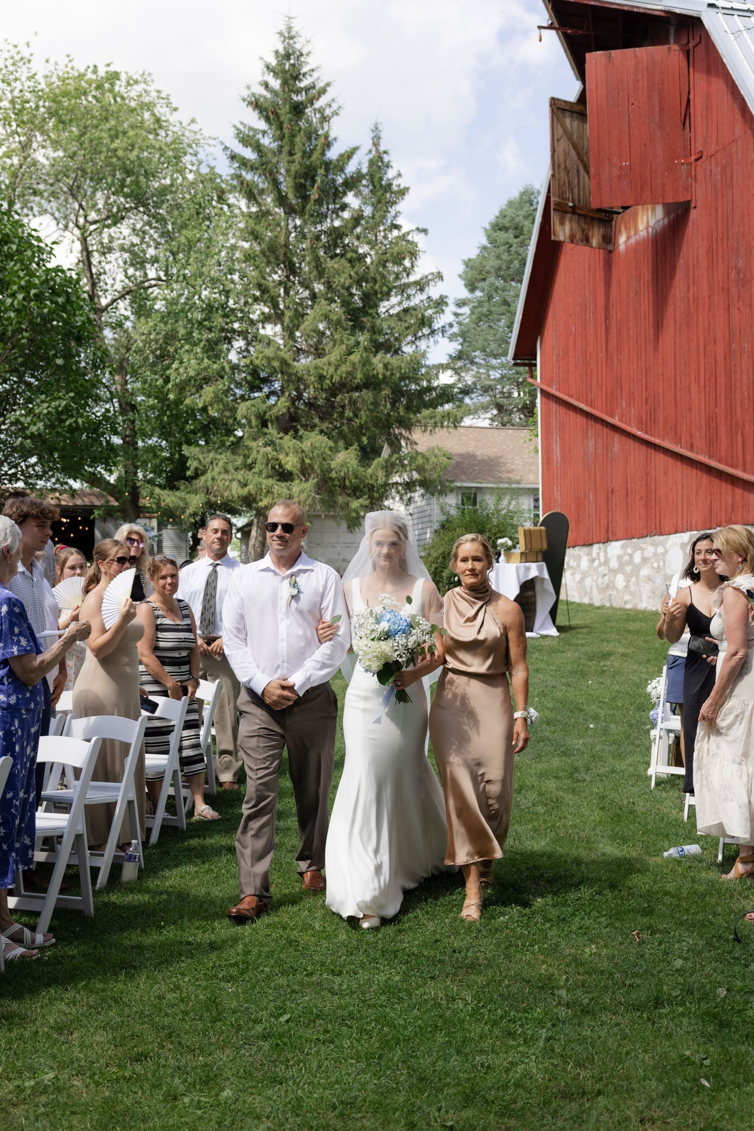 Wedding ceremony processional on grass with red barn and guests in attendance.
