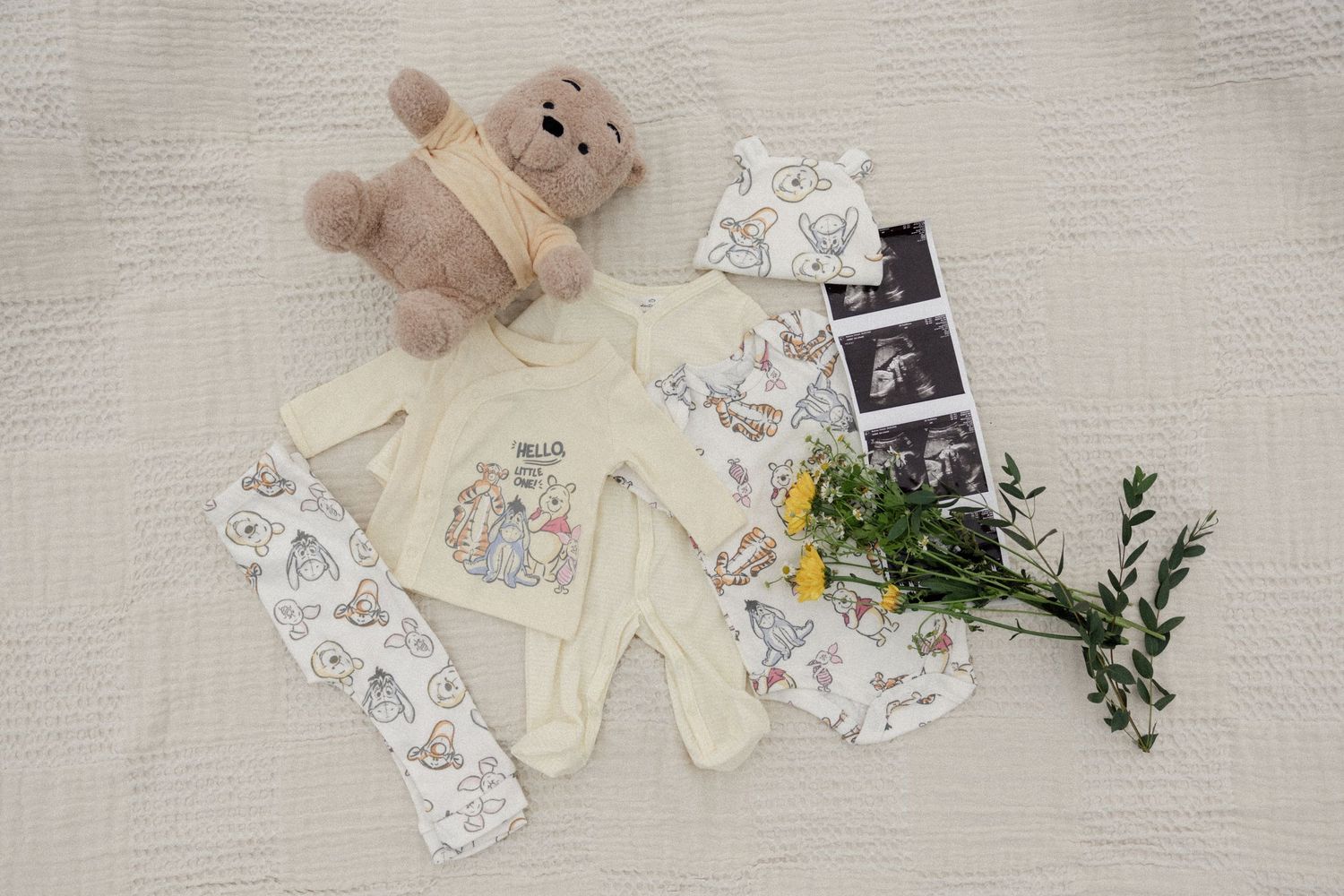 Pregnancy announcement flatlay with baby clothes, ultrasound photos, teddy bear, and floral accents on light fabric.