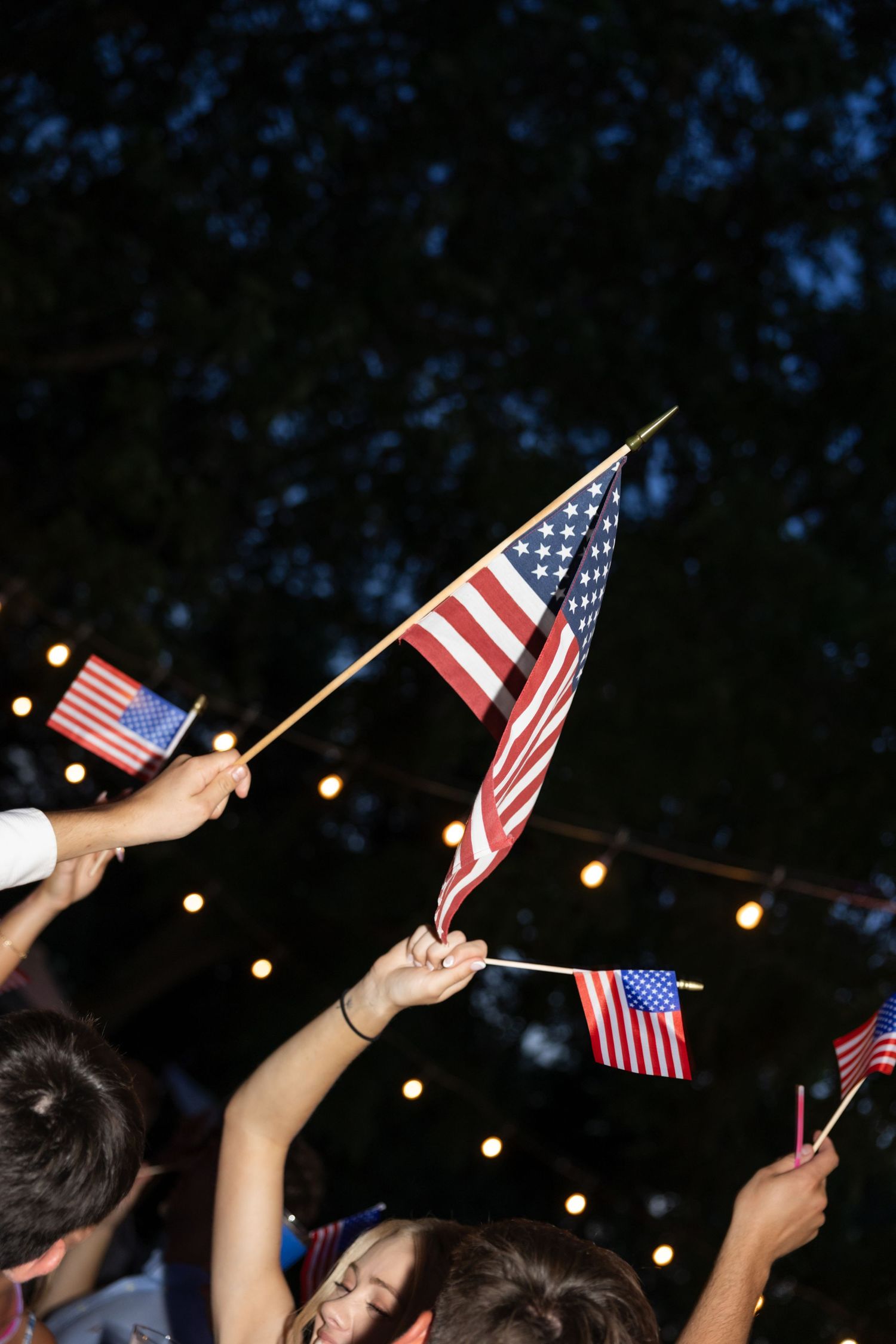 Hands raised holding small American flags against dark sky with string lights.