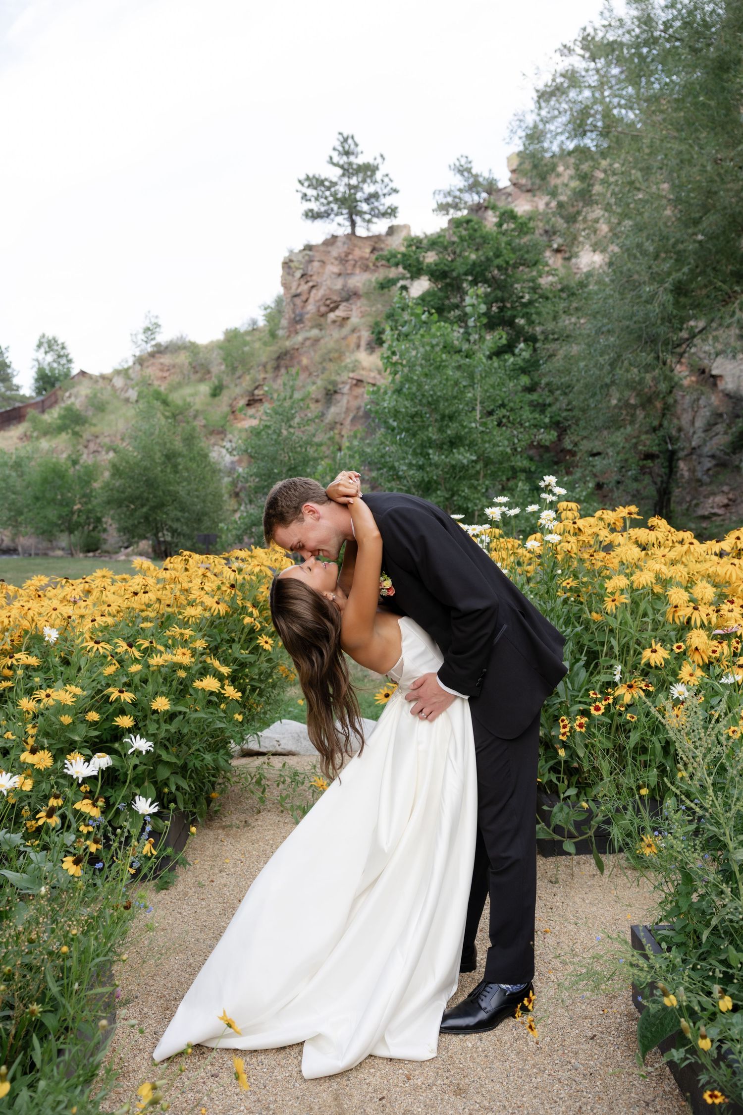 A dramatic dip kiss between newlyweds surrounded by yellow wildflowers on a garden path.
