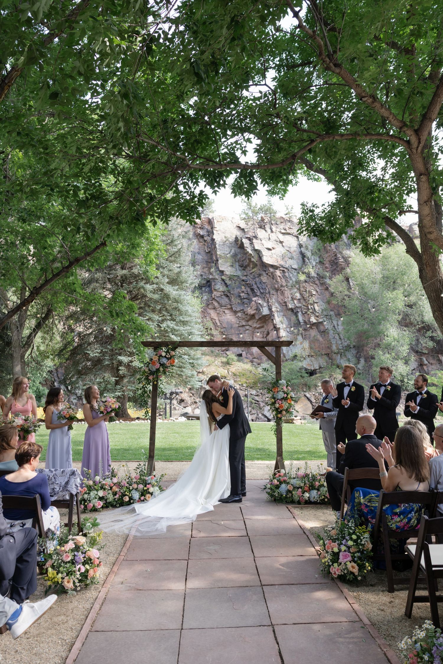 An outdoor wedding ceremony with guests seated along a flower-lined aisle beneath towering trees and cliffs.