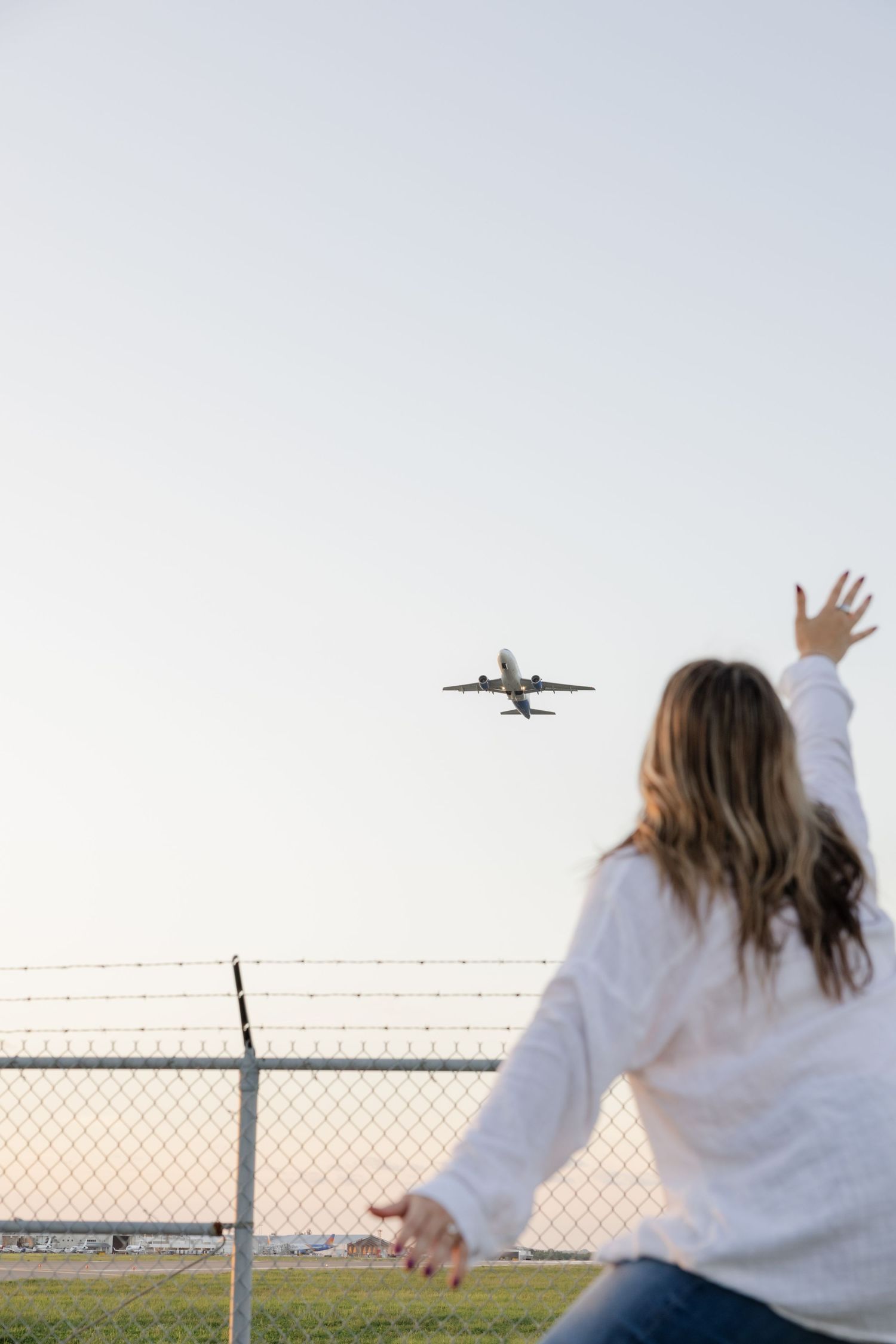Group watching and waving at airplane taking off from behind chain link fence at airport.