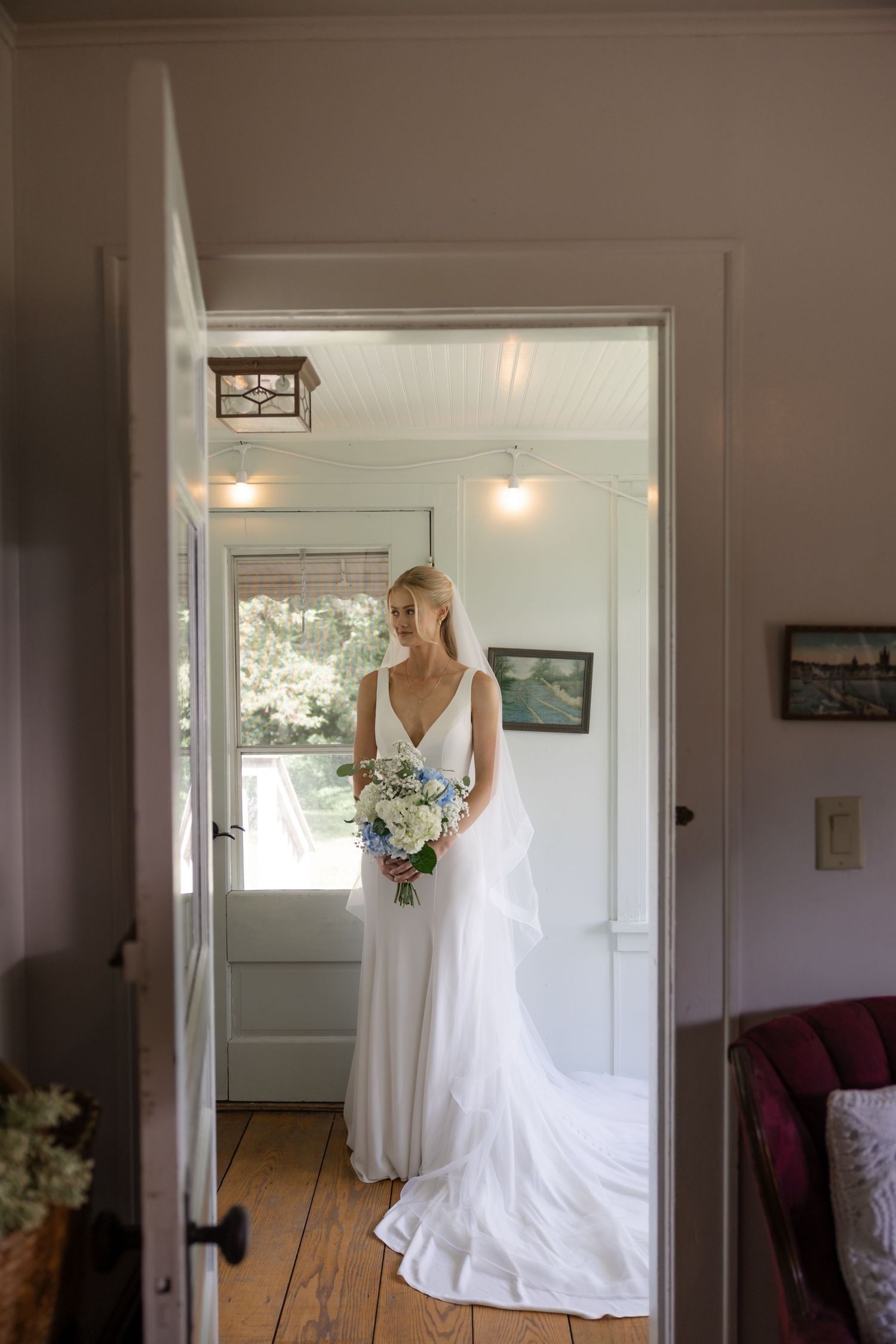 White wedding dress hangs in doorway of sunlit room with wooden floors.