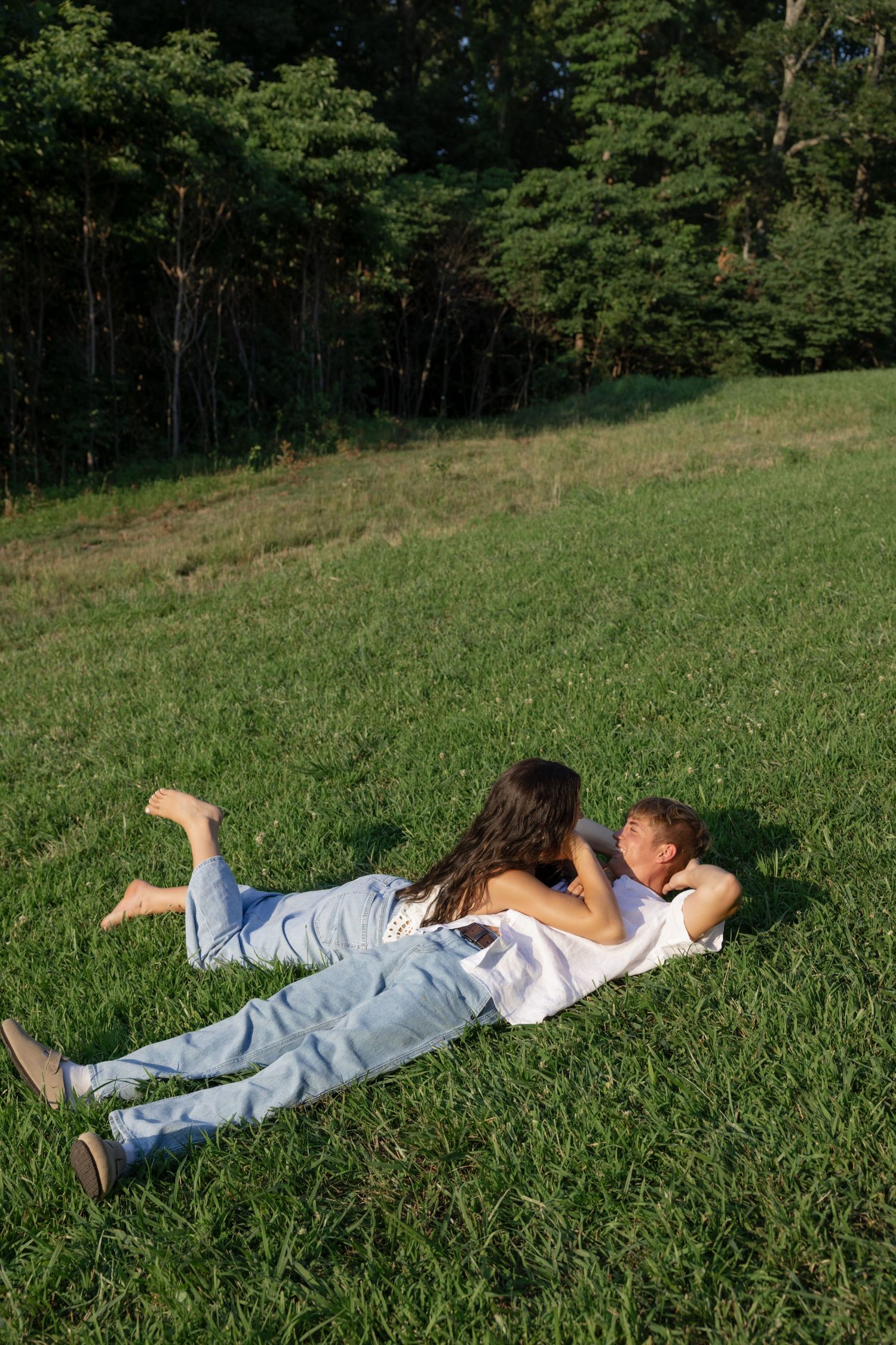 A couple relaxes together on green grass near a wooded area on a sunny day.