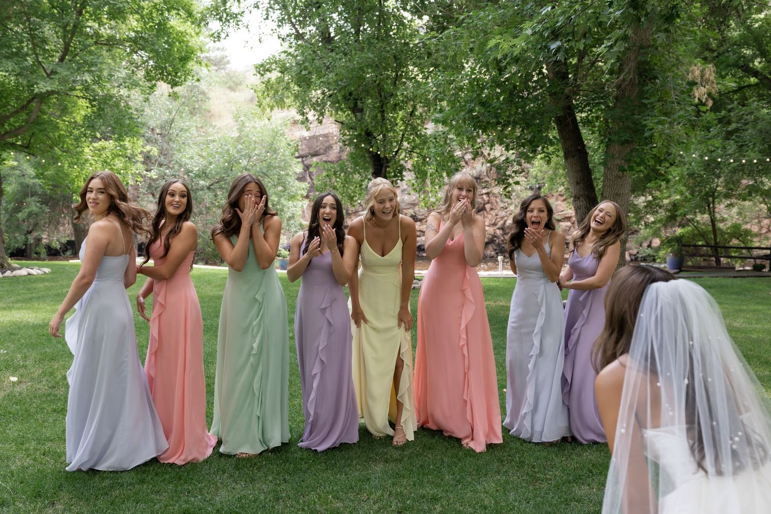 Row of bridesmaids in pastel dresses sitting together in a garden setting.