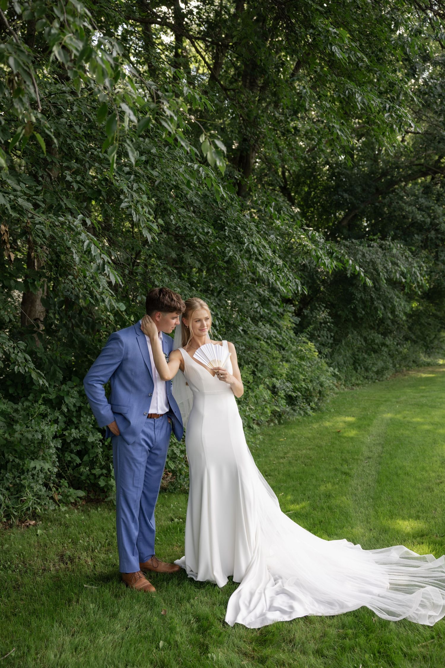 Newlyweds in blue and white share tender moments on grassy lawn surrounded by trees.