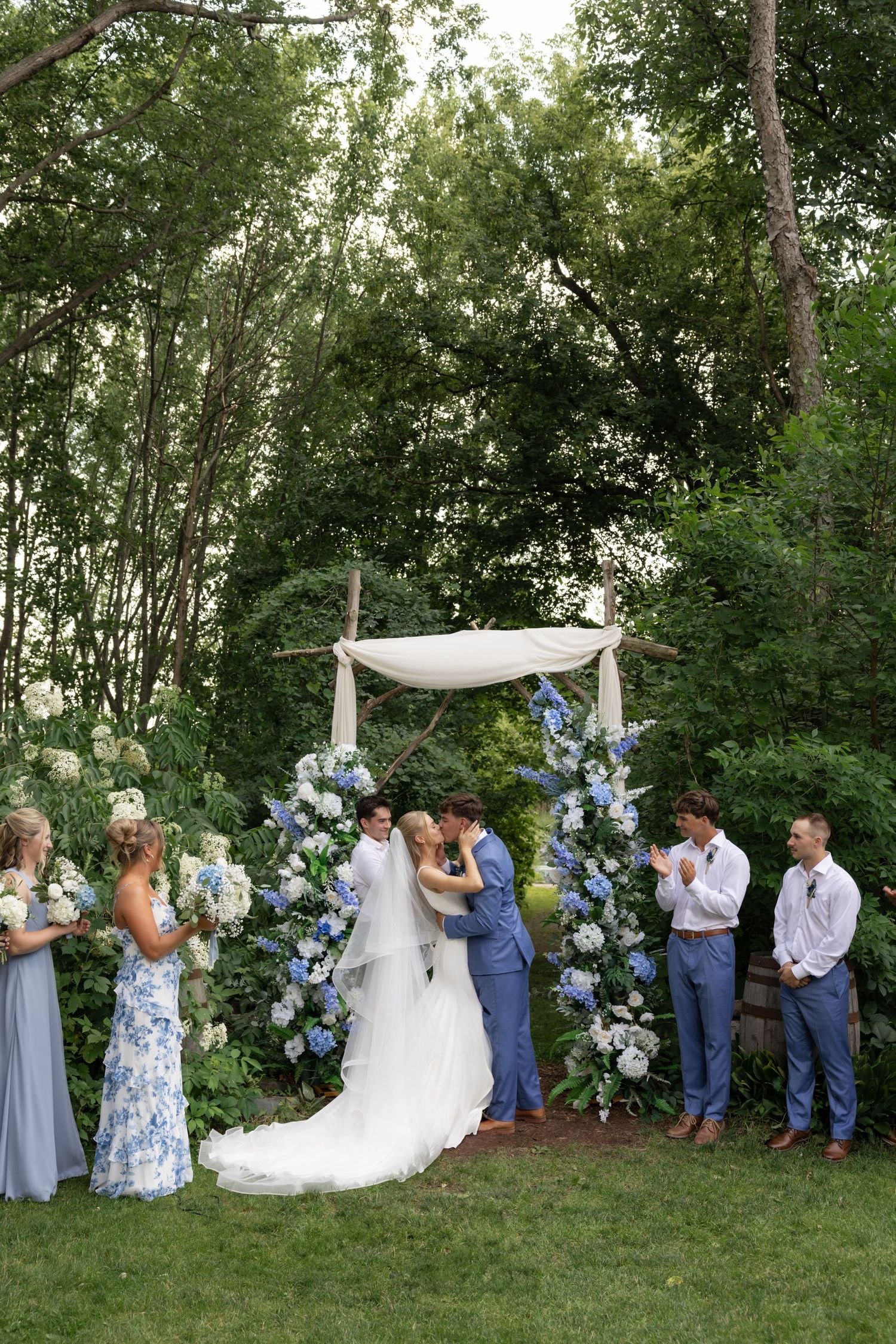 Wedding party lined up outdoors with white and blue floral decorations against a backdrop of green trees.