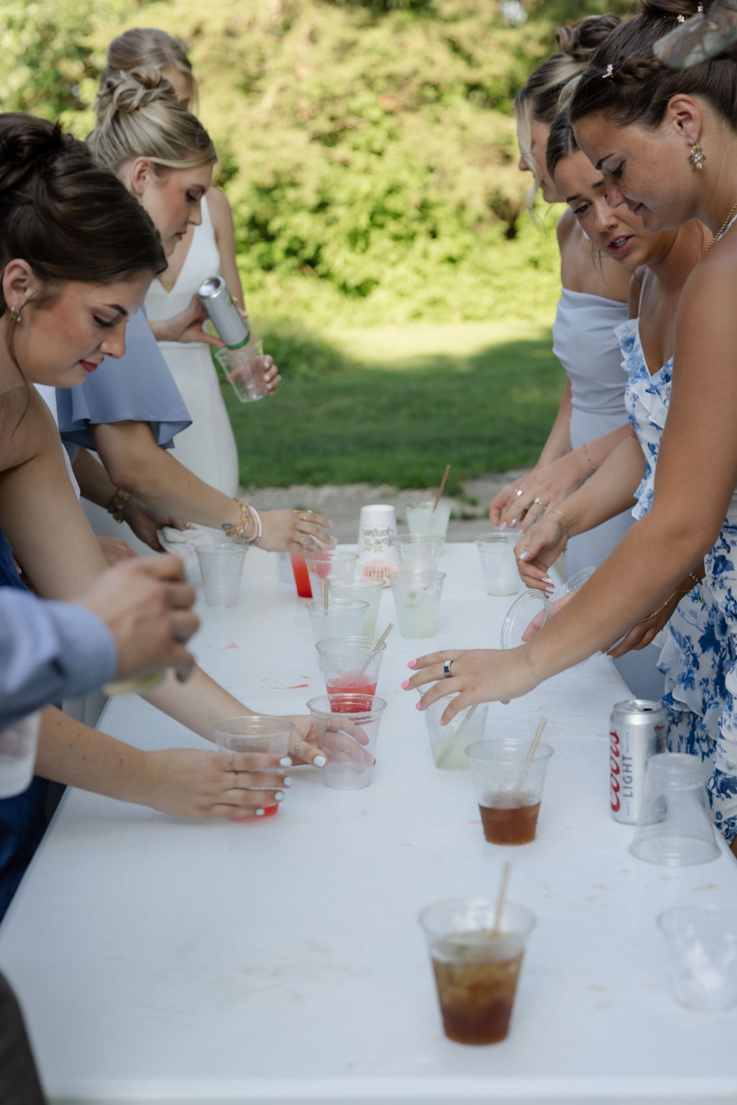 Wedding guests gather around a white table preparing drinks at an outdoor reception.