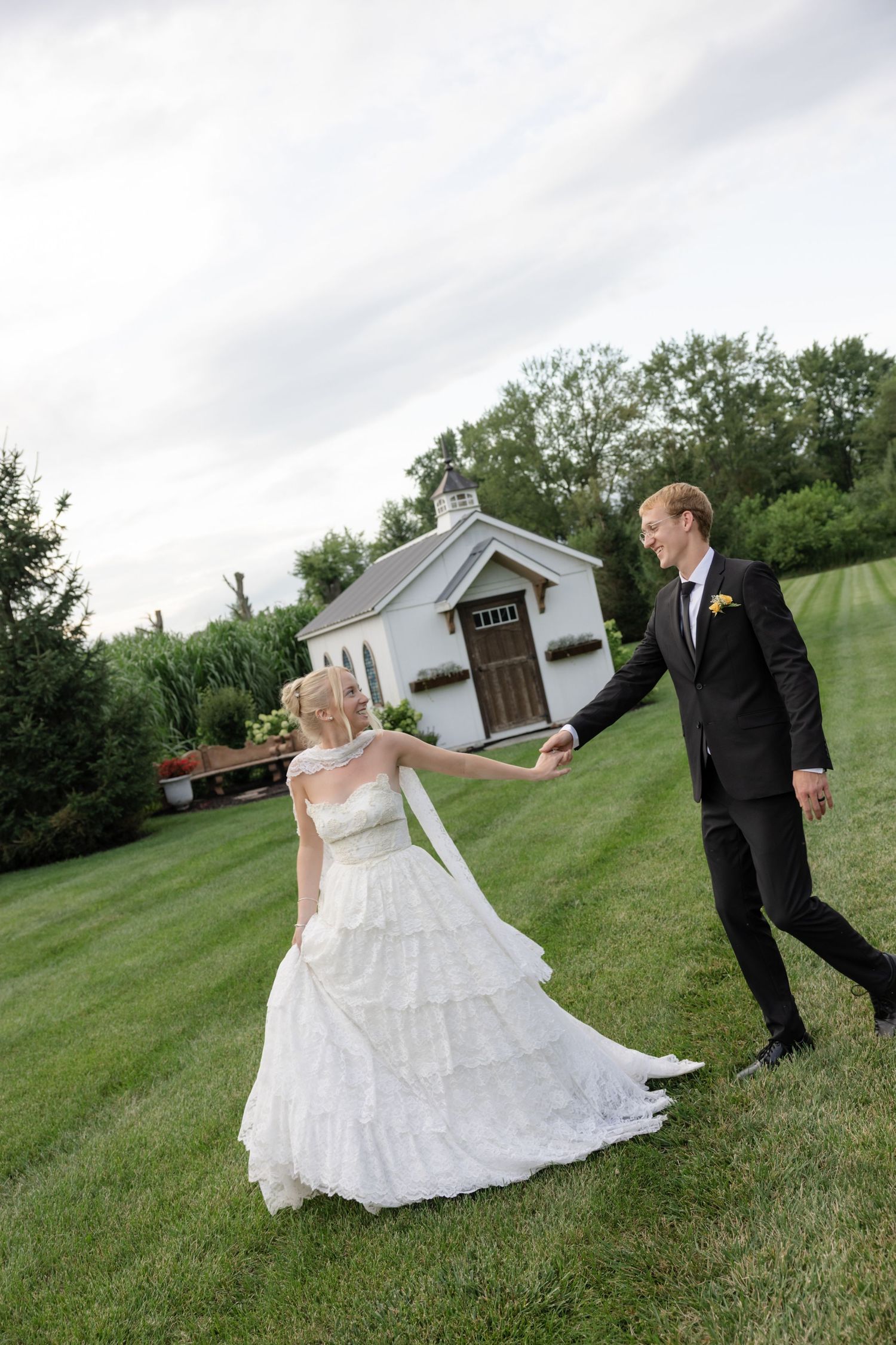 Playful wedding couple poses across grass field near white buildings.