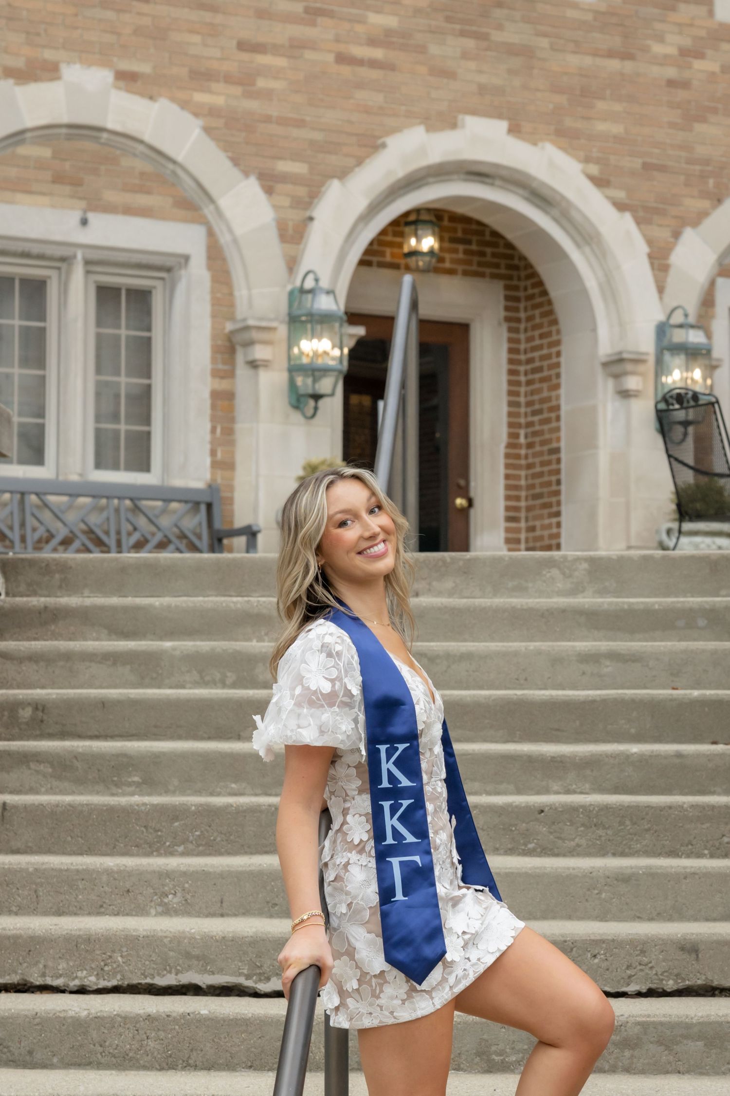 Student in floral dress and blue sorority sash poses on steps of brick building.