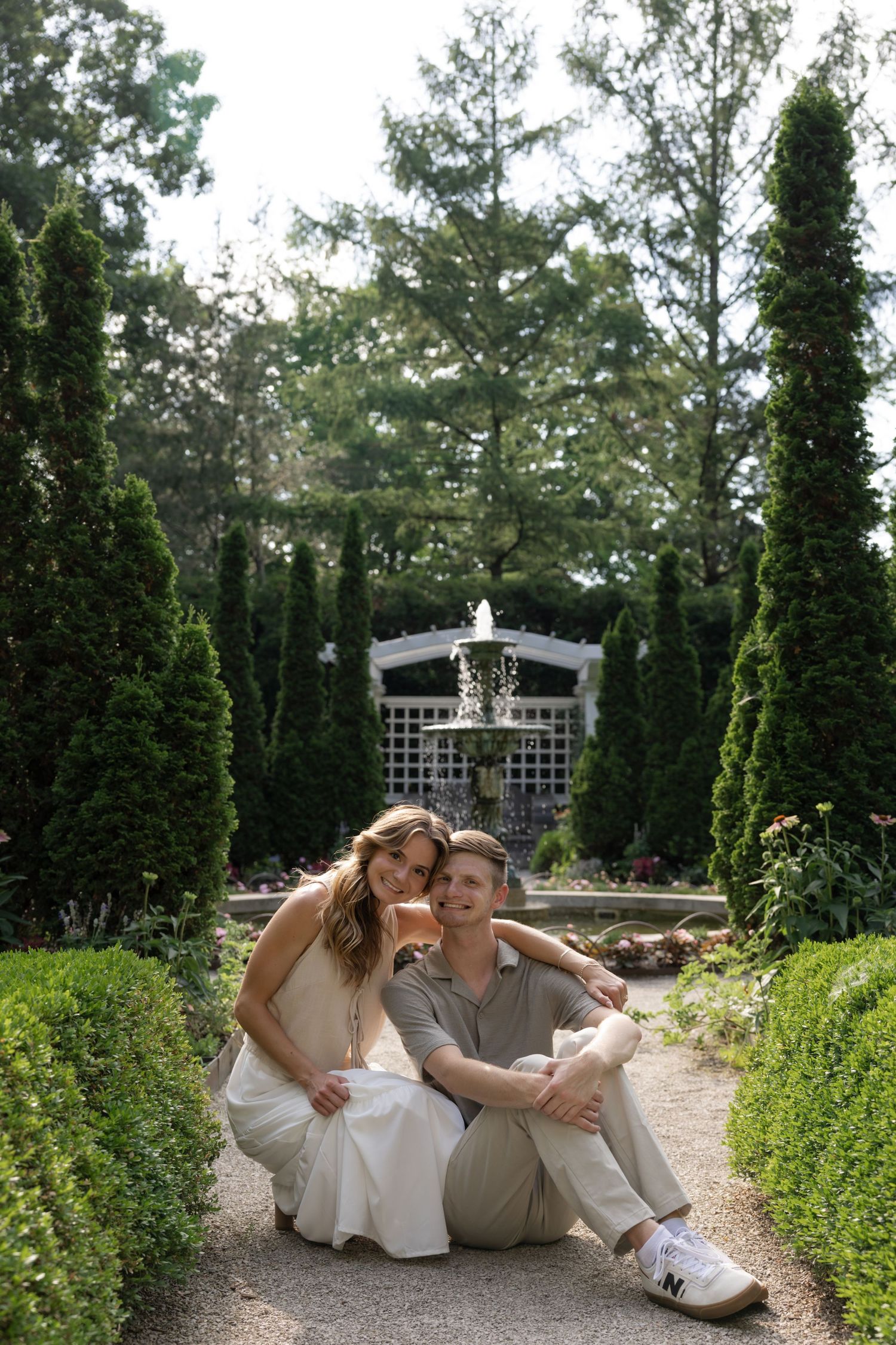A couple relaxes in a formal garden with topiary and a decorative fountain structure.
