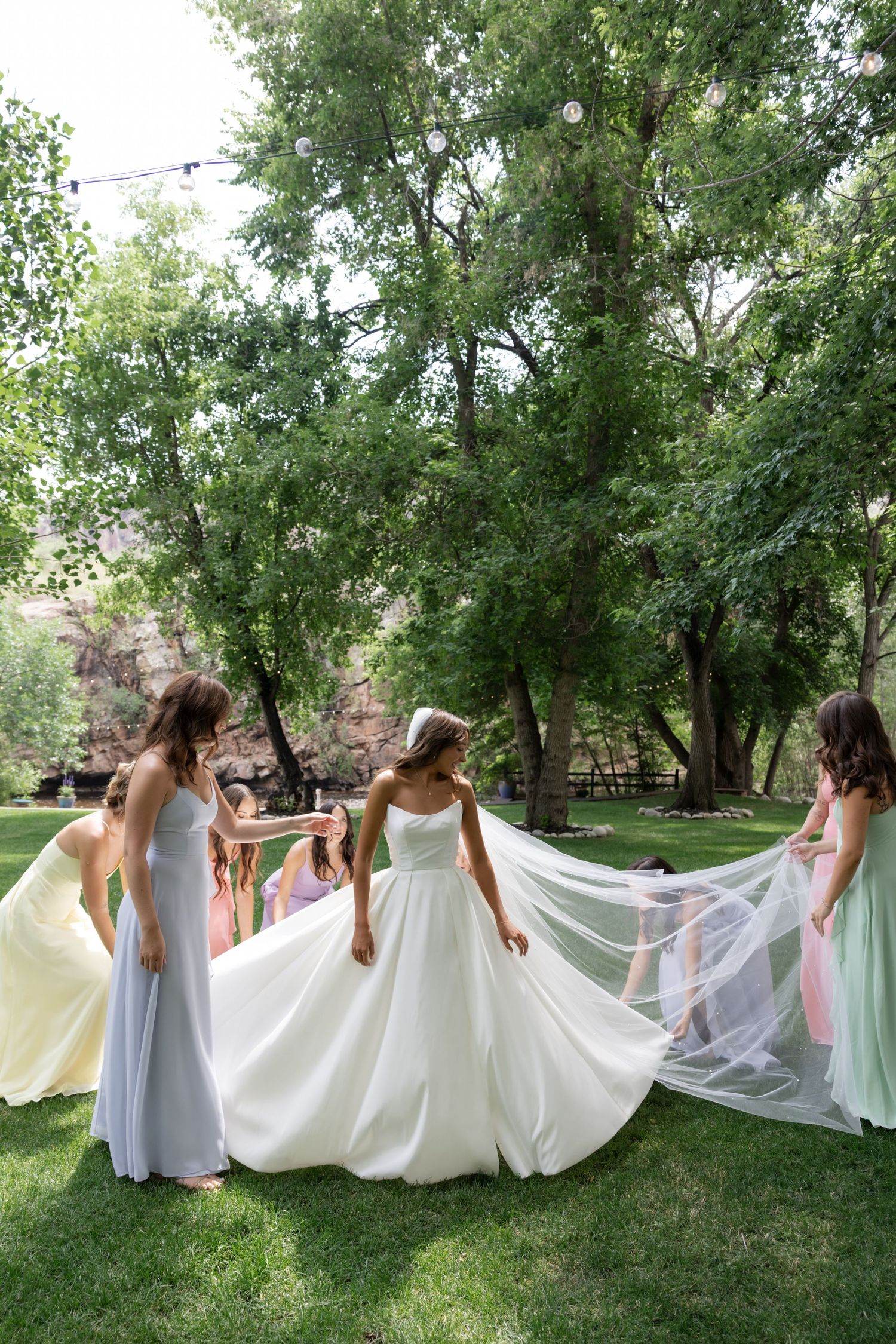 Wedding party spreads out white dress train in a line across green lawn.