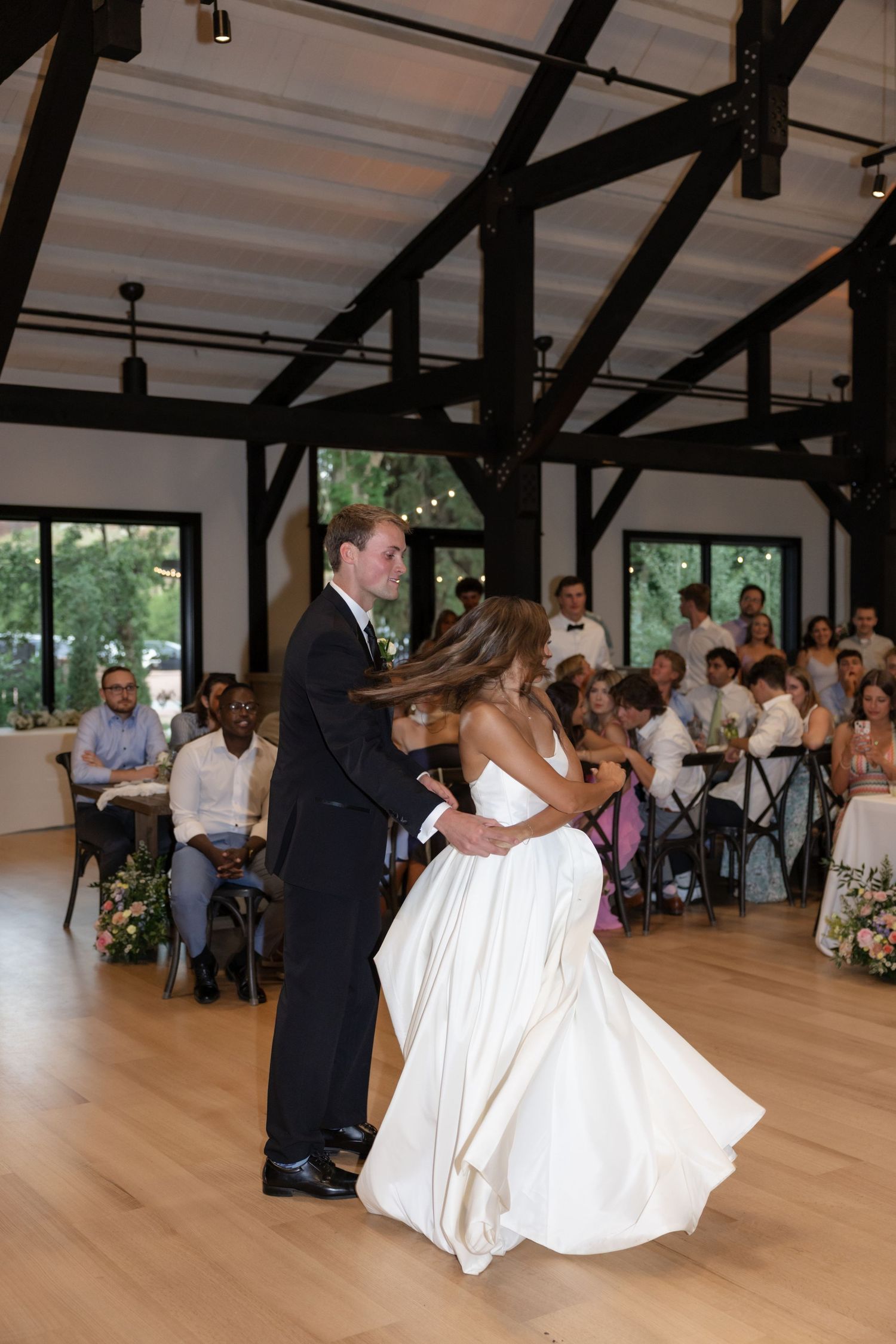 Bride in flowing white gown dances with groom in elegant barn venue with exposed beams.
