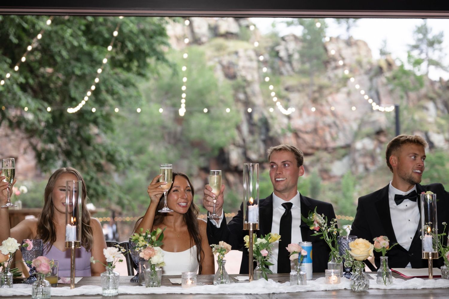 Wedding party seated at long tables during outdoor reception with string lights.