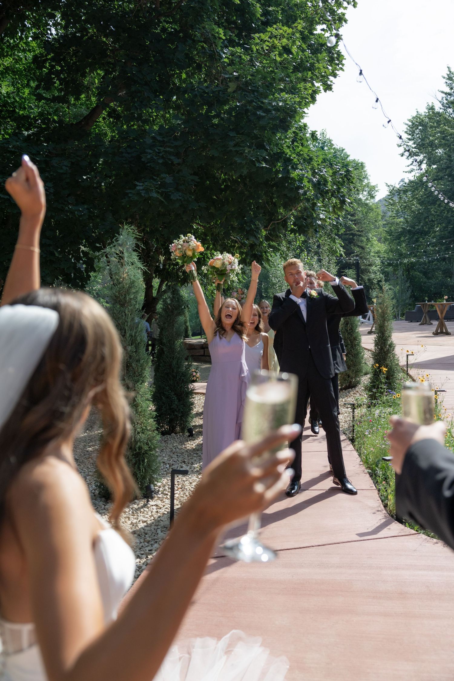 Wedding guests raise champagne glasses in celebration on an outdoor walkway.
