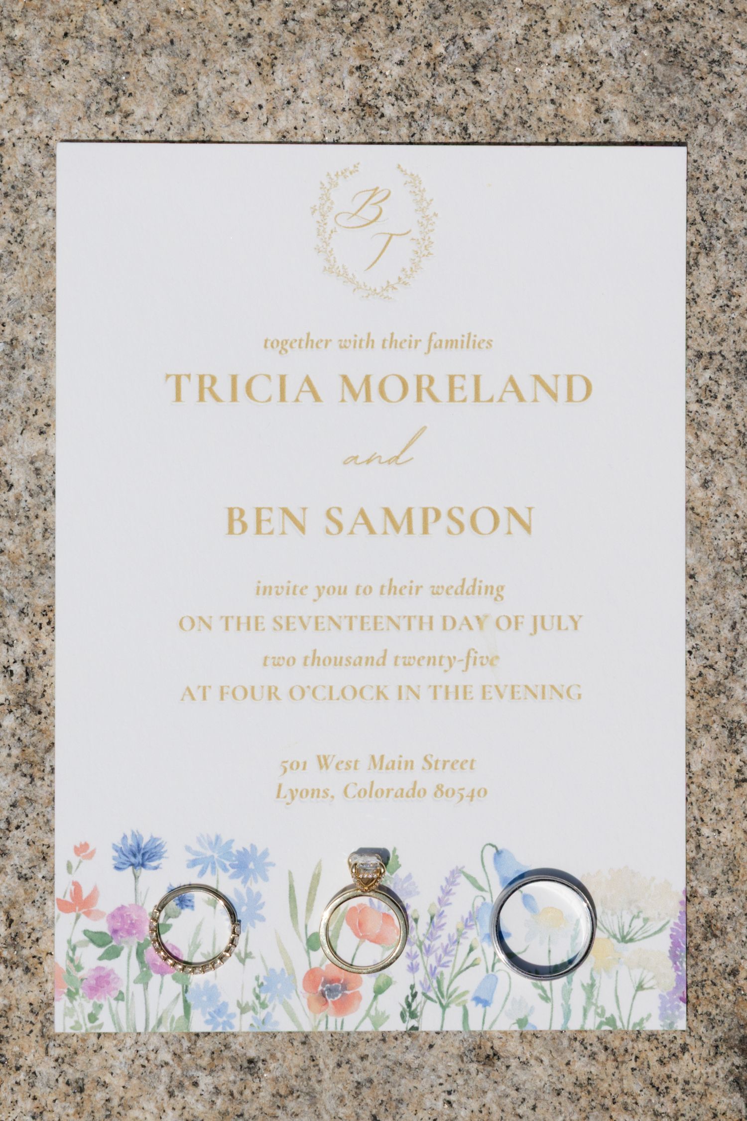Elegant wedding invitation with watercolor floral design and gold text on stone background.