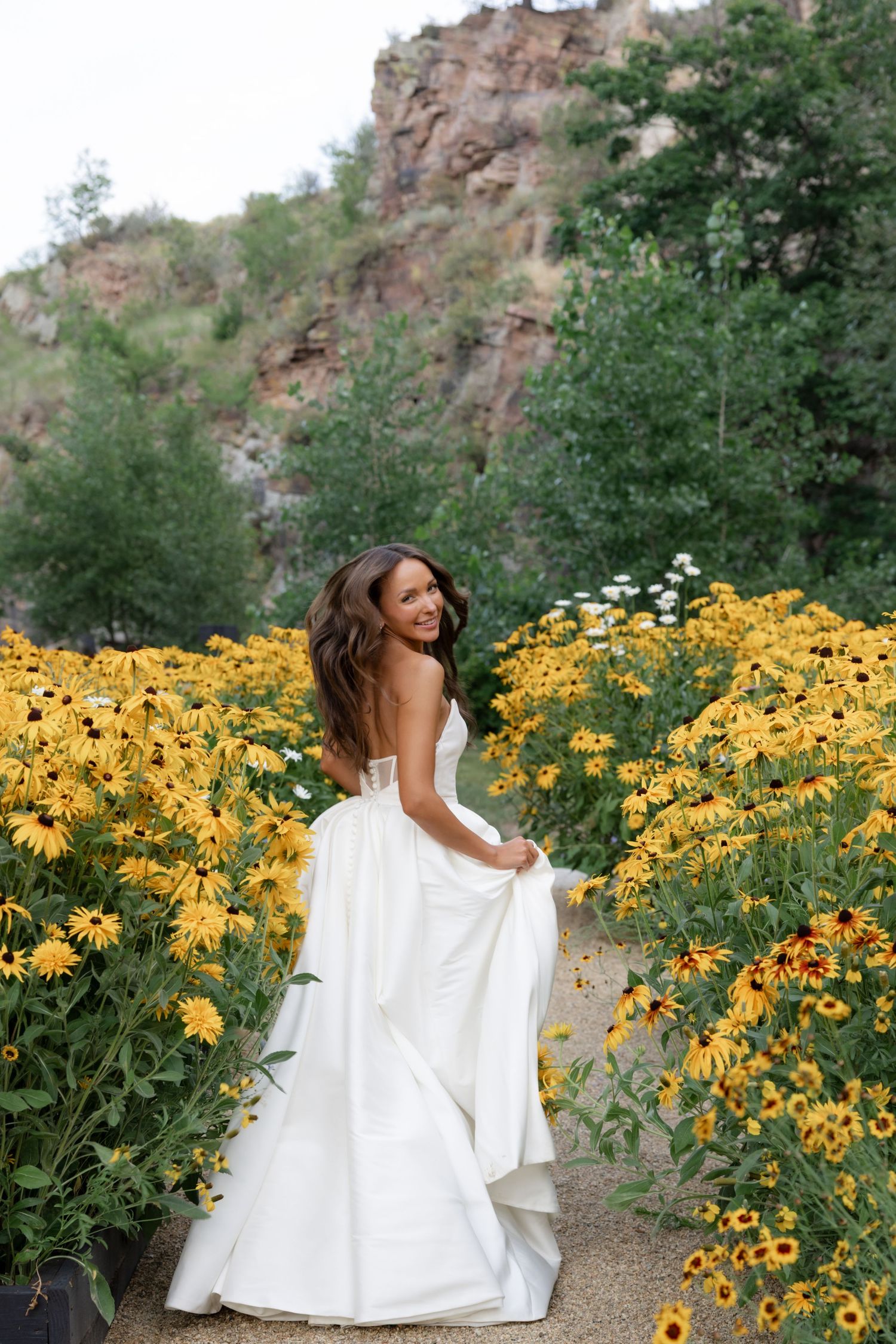 A bride in a flowing white gown poses among vibrant yellow wildflowers with rocky cliffs behind her.
