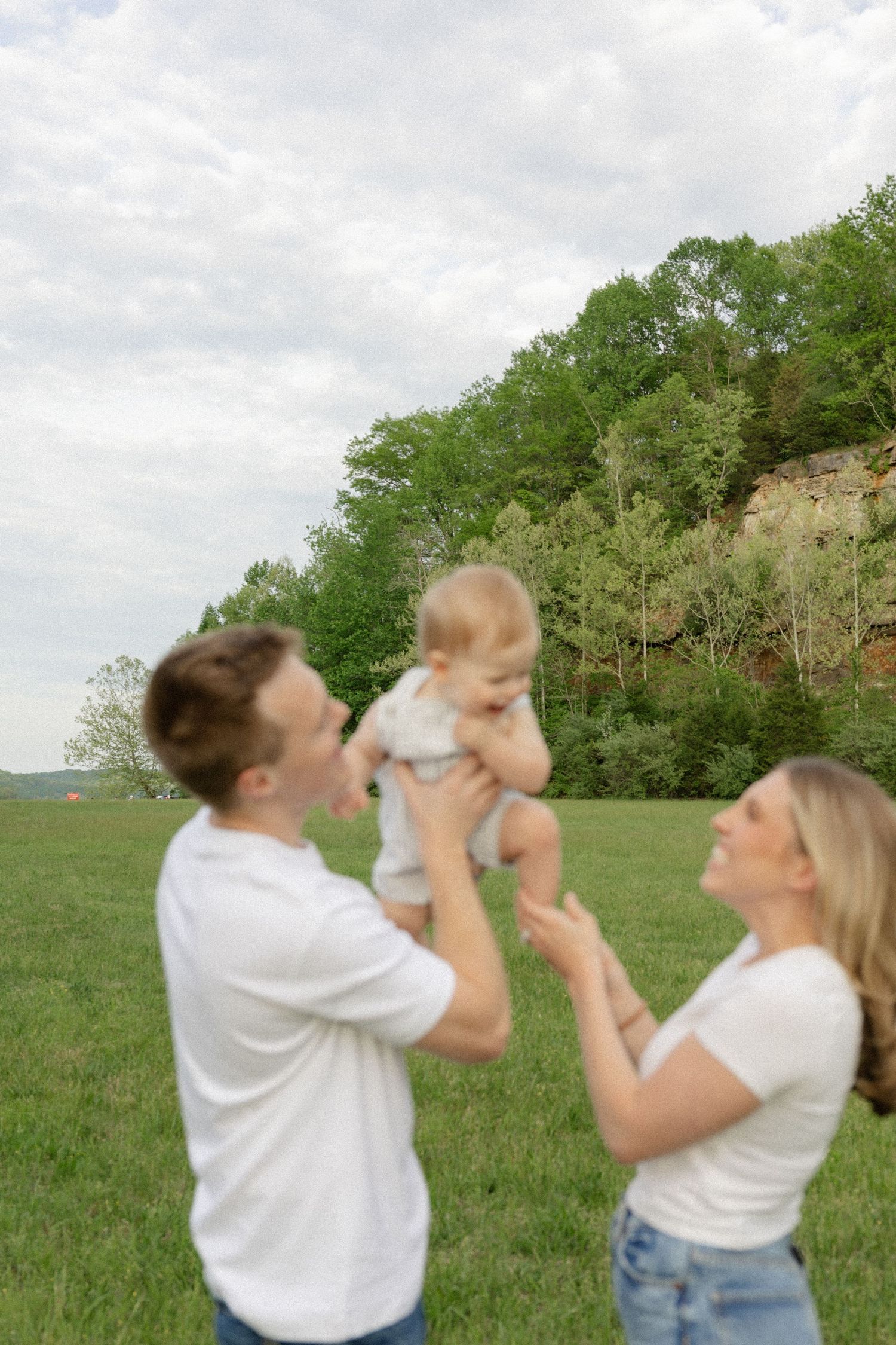 Happy couple lifts baby in the air while playing in a scenic meadow.