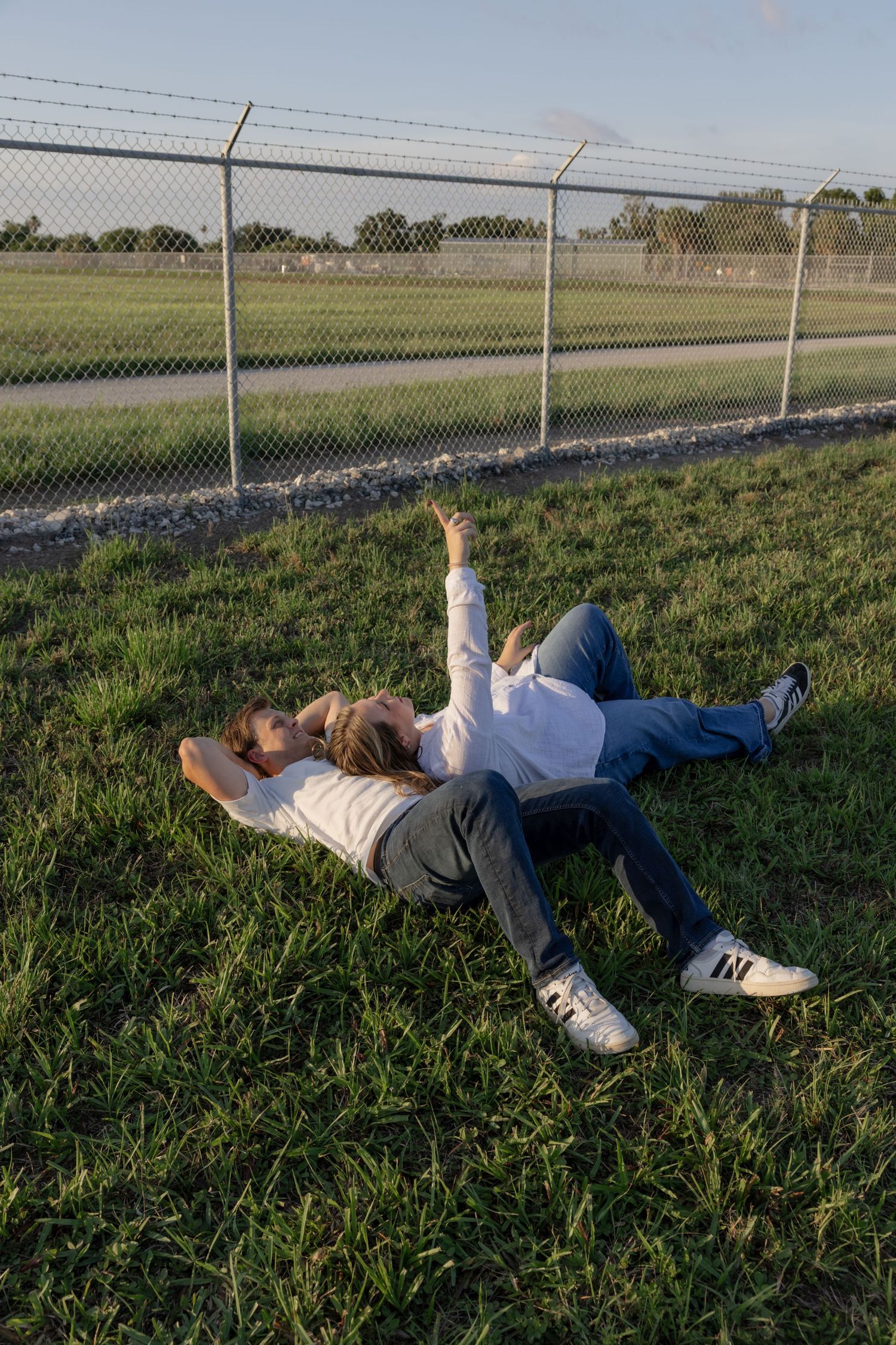 Friends lying on grass waving at planes passing overhead near airport fence.