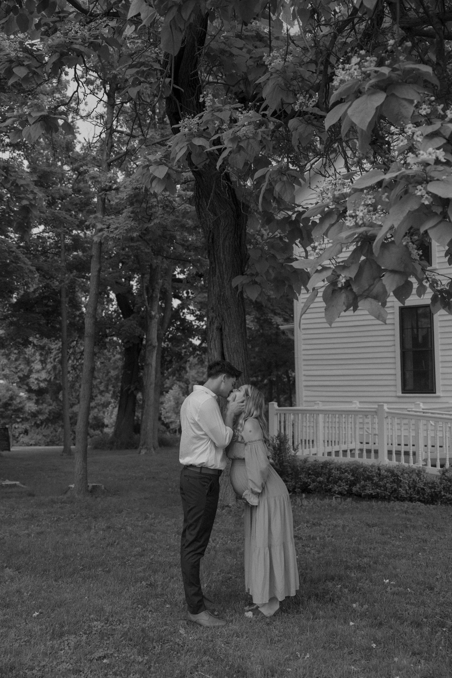 A romantic black and white portrait of a couple standing near a white colonial house.