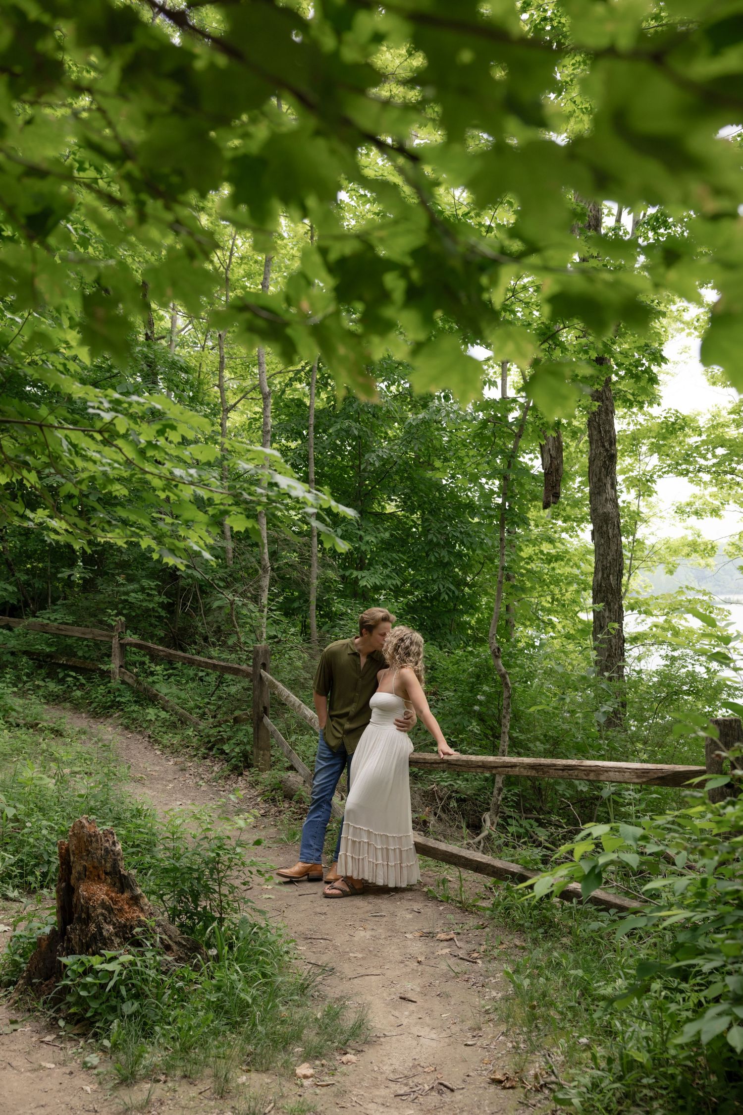 Two people walking together on a wooded dirt path surrounded by lush green foliage.