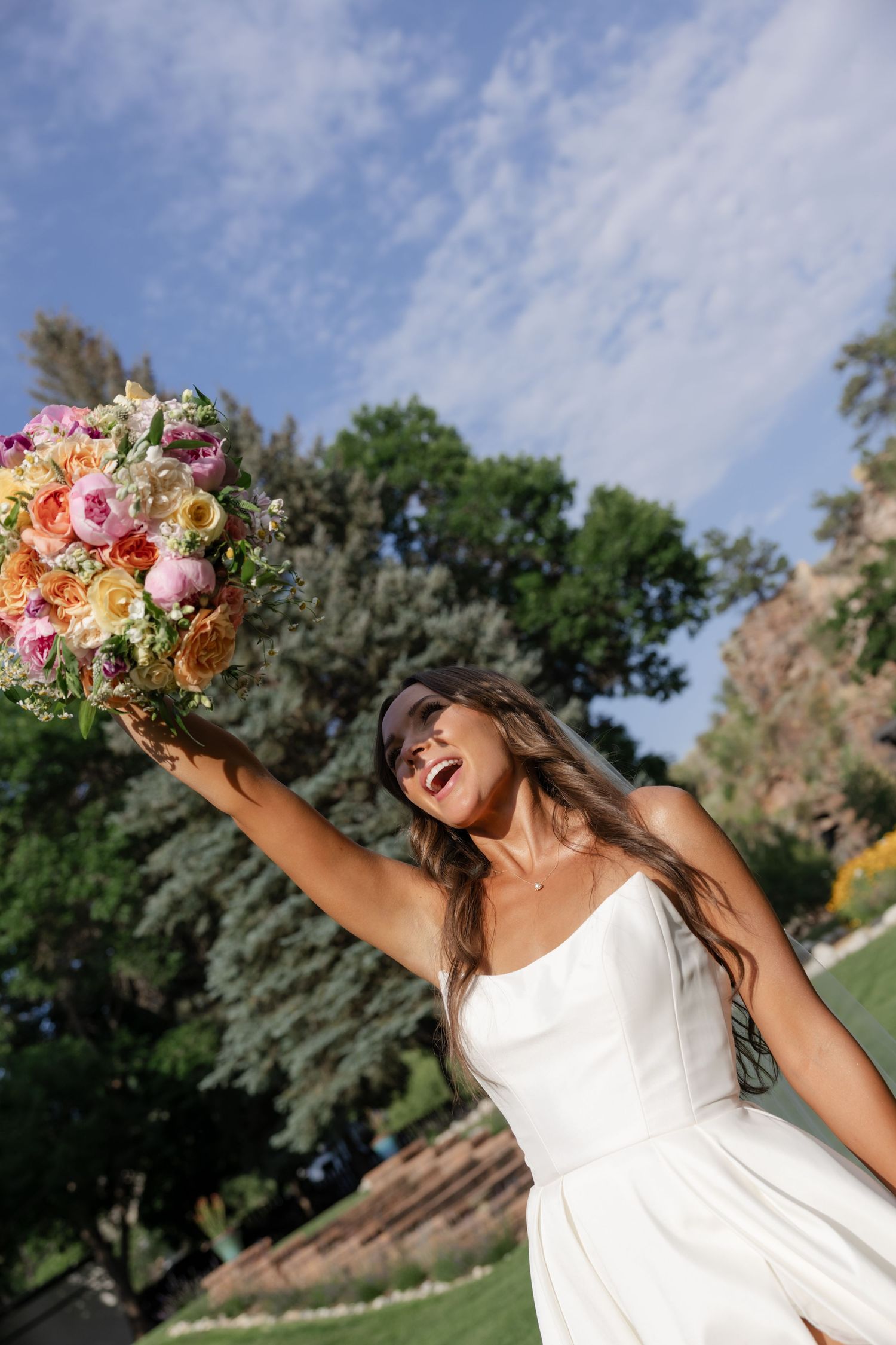 Joyful bride raises colorful bouquet overhead against blue sky and mountain landscape.