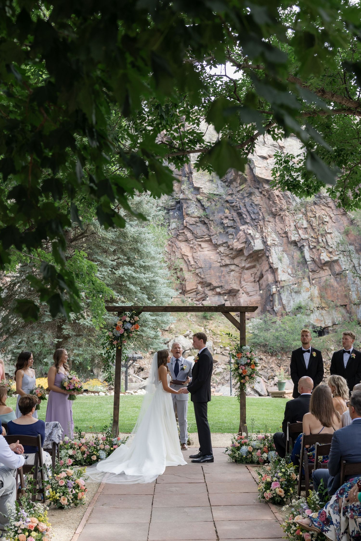 Wedding ceremony setup with wooden arch and floral arrangements against a dramatic rock wall backdrop.