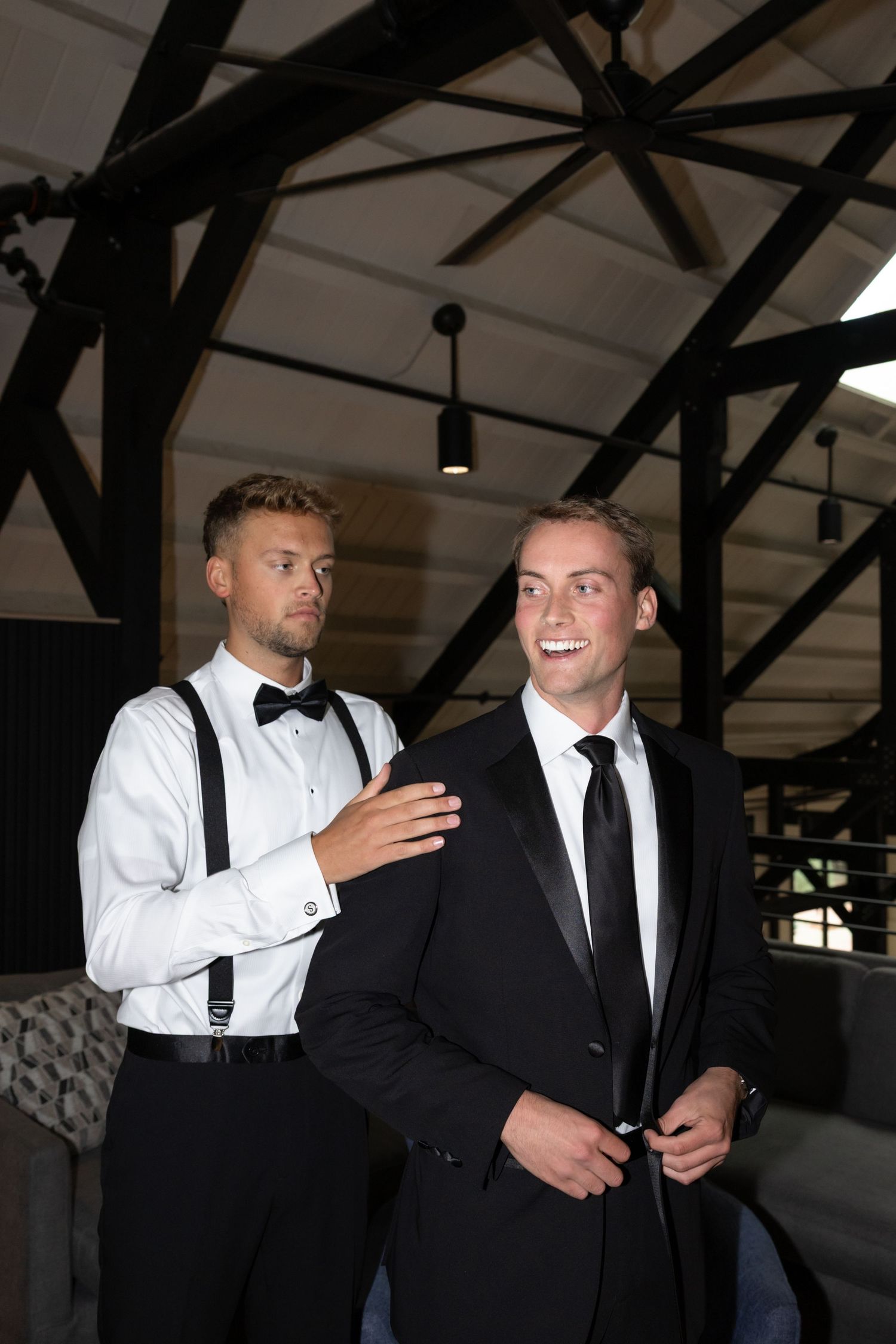 Two men in formal attire share a moment under architectural wooden beams.