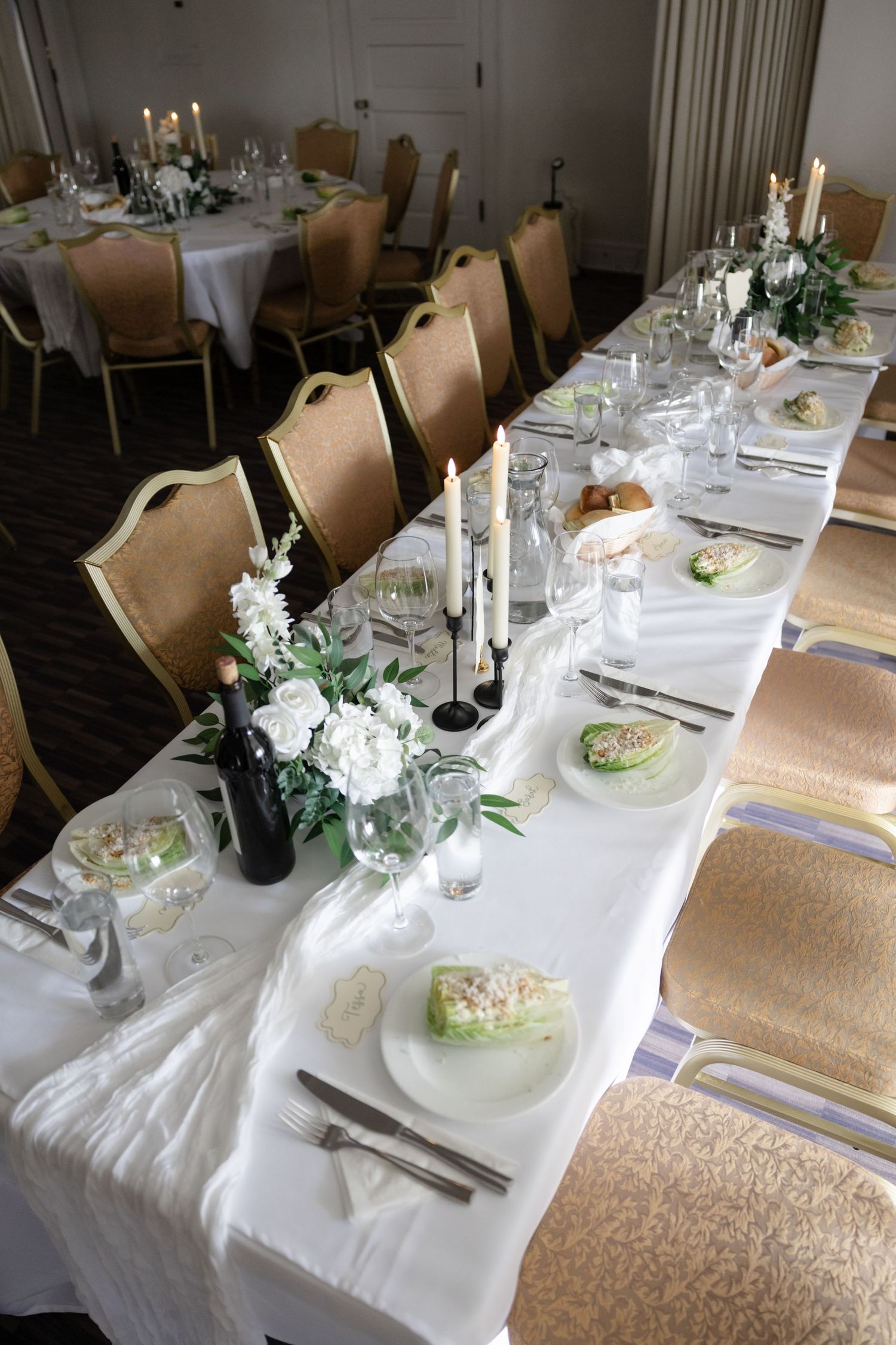 An elegant wedding reception table setting with white linens, candles, and greenery centerpieces.