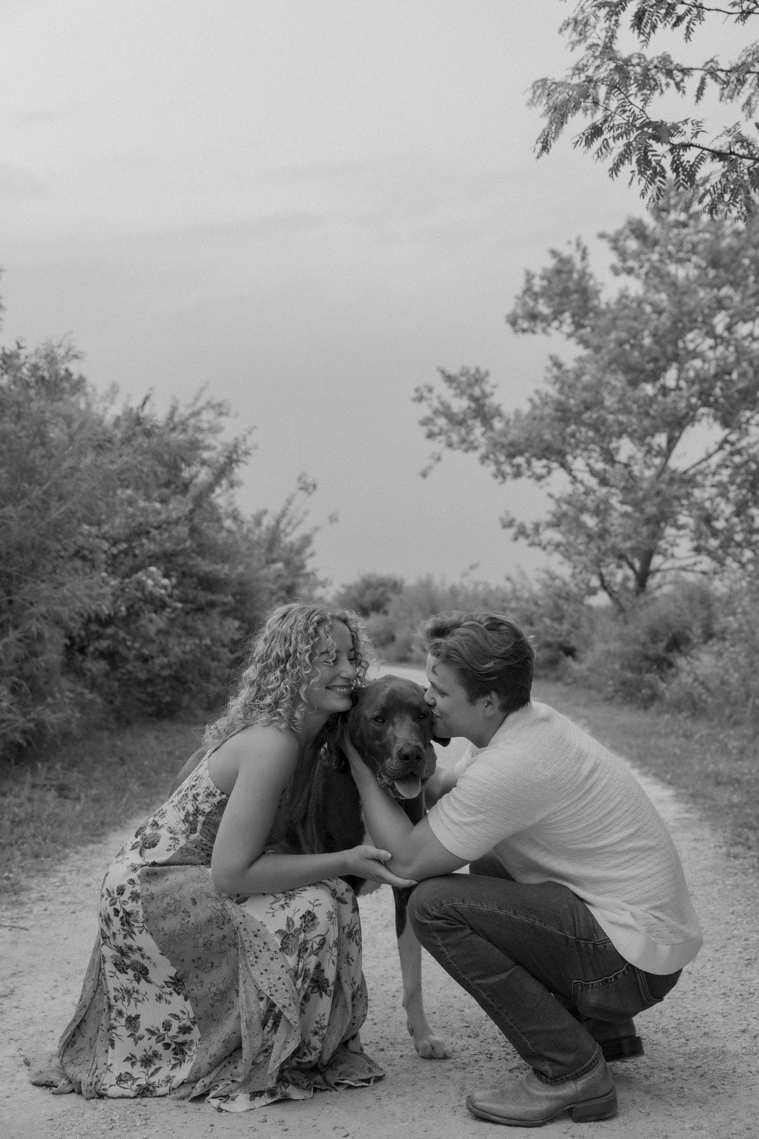 A romantic couple sits close together on a dirt path surrounded by greenery.