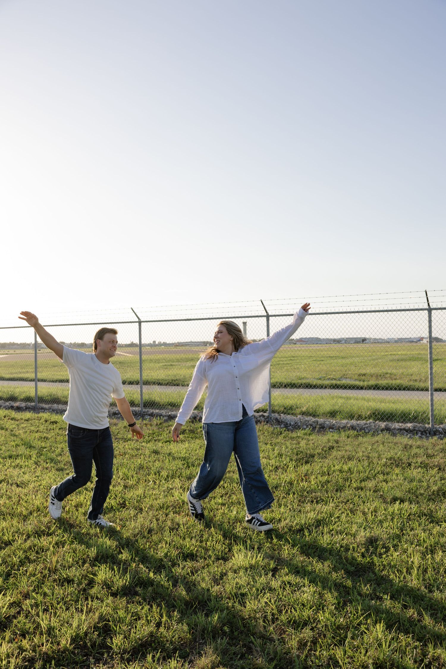 A couple running playfully through a grassy field next to a chain-link fence.